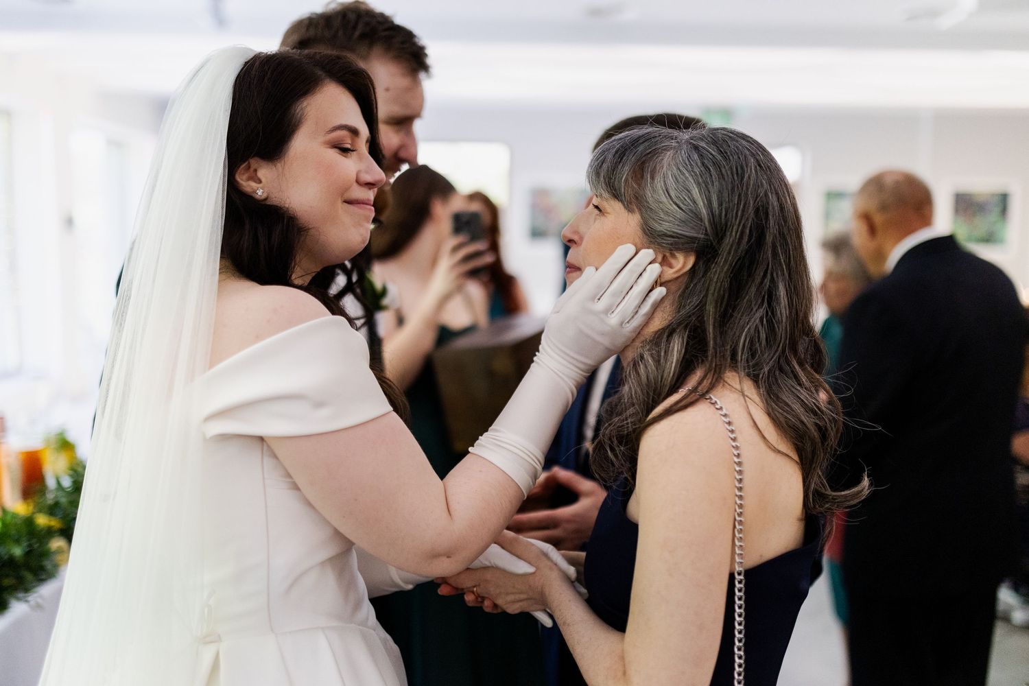 An emotional moment between loved ones at a wedding reception with guests in the background.