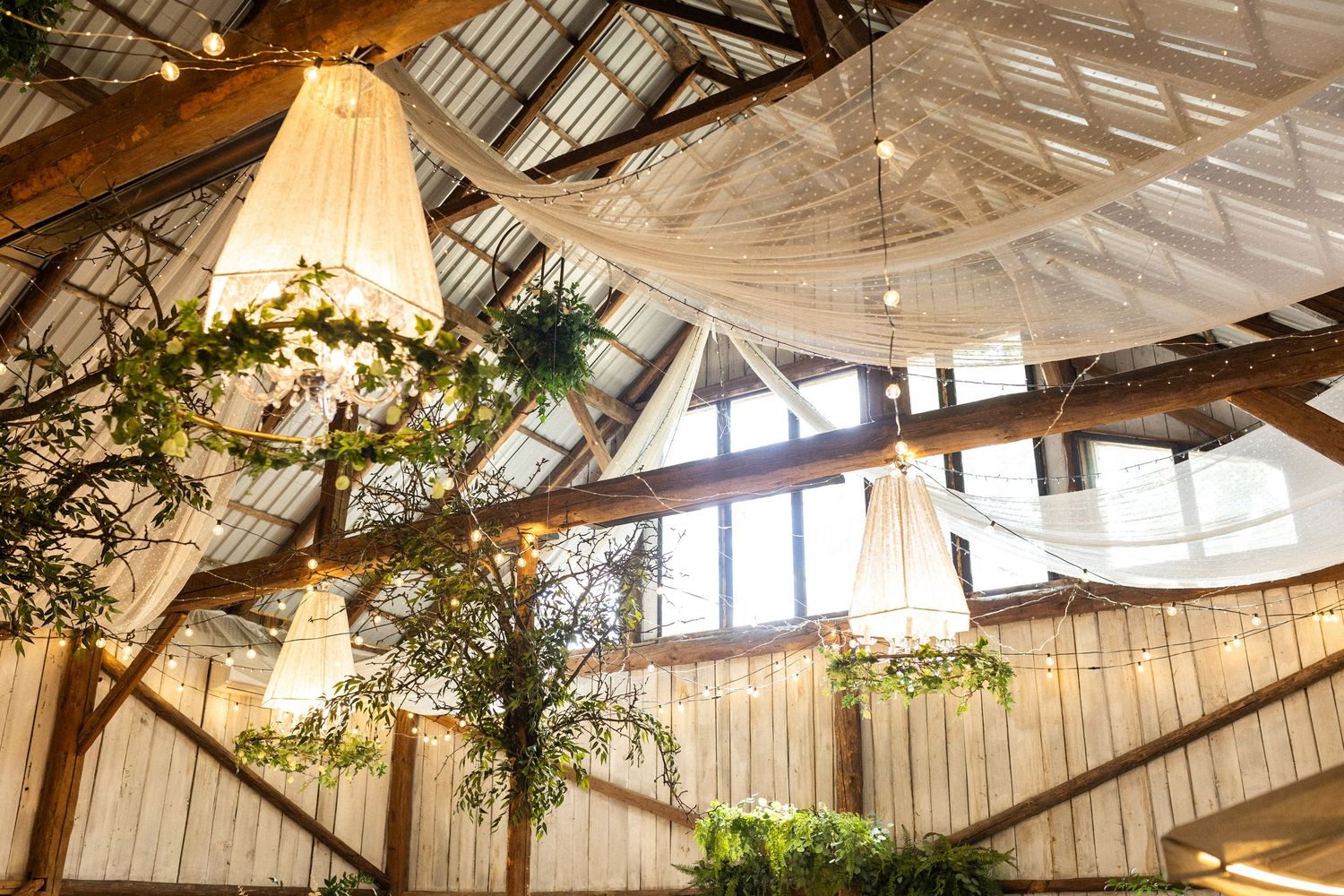 Large rustic chandelier hangs from exposed wooden beams in barn venue with draped fabric and greenery decorations.