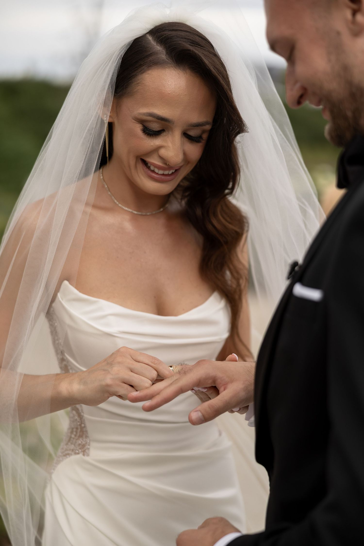 Smiling bride in white gown and veil placing wedding ring on grooms finger during outdoor ceremony.