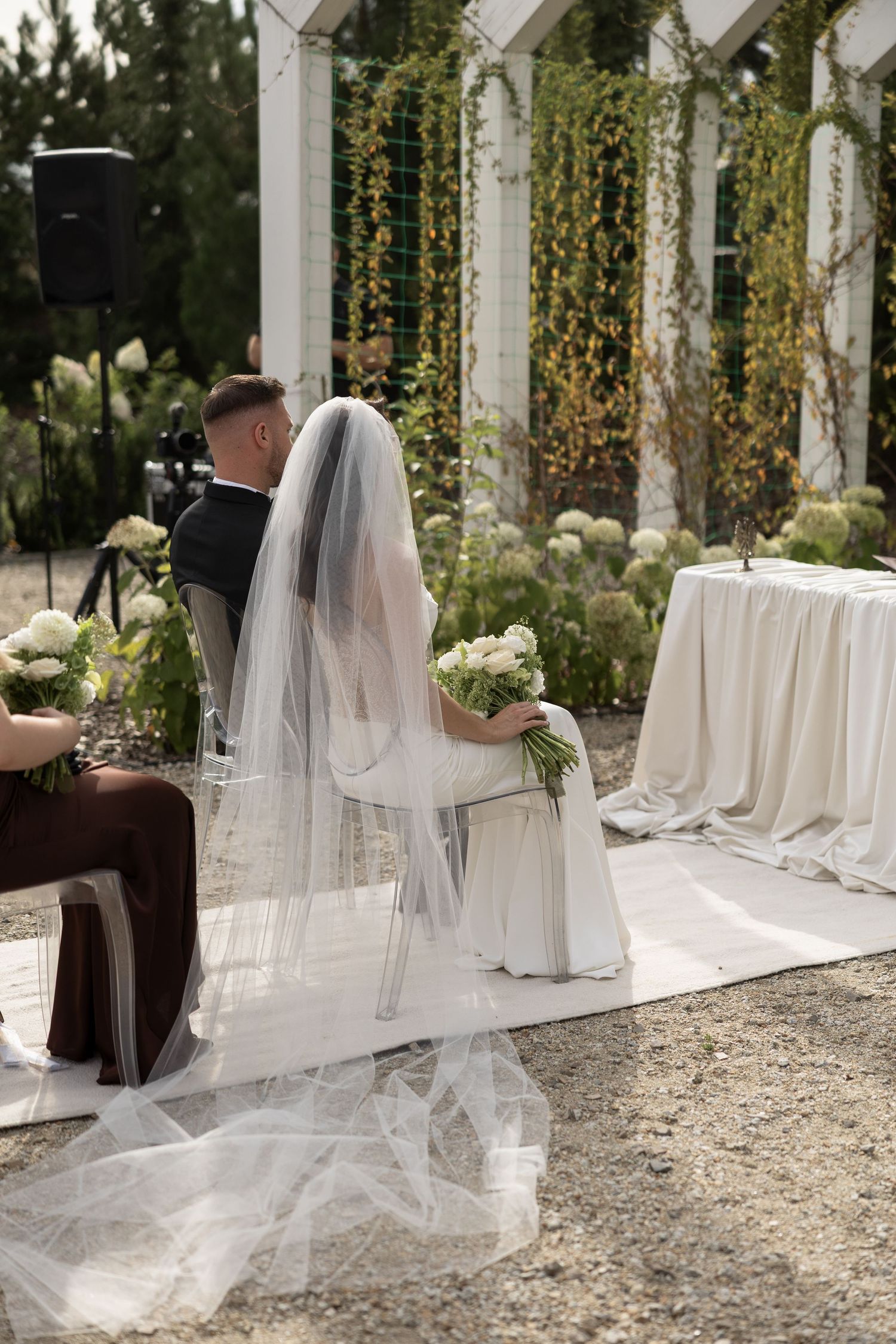 Bride and groom seated at outdoor wedding ceremony with white floral arrangements and lush green pergola backdrop.