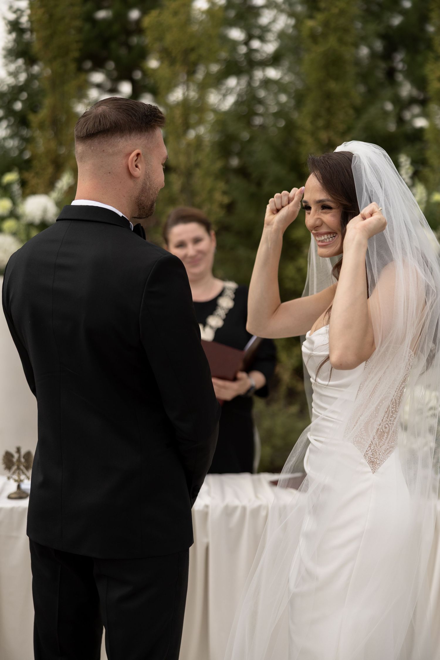 Bride smiling and lifting her veil during outdoor wedding ceremony while groom stands before her.