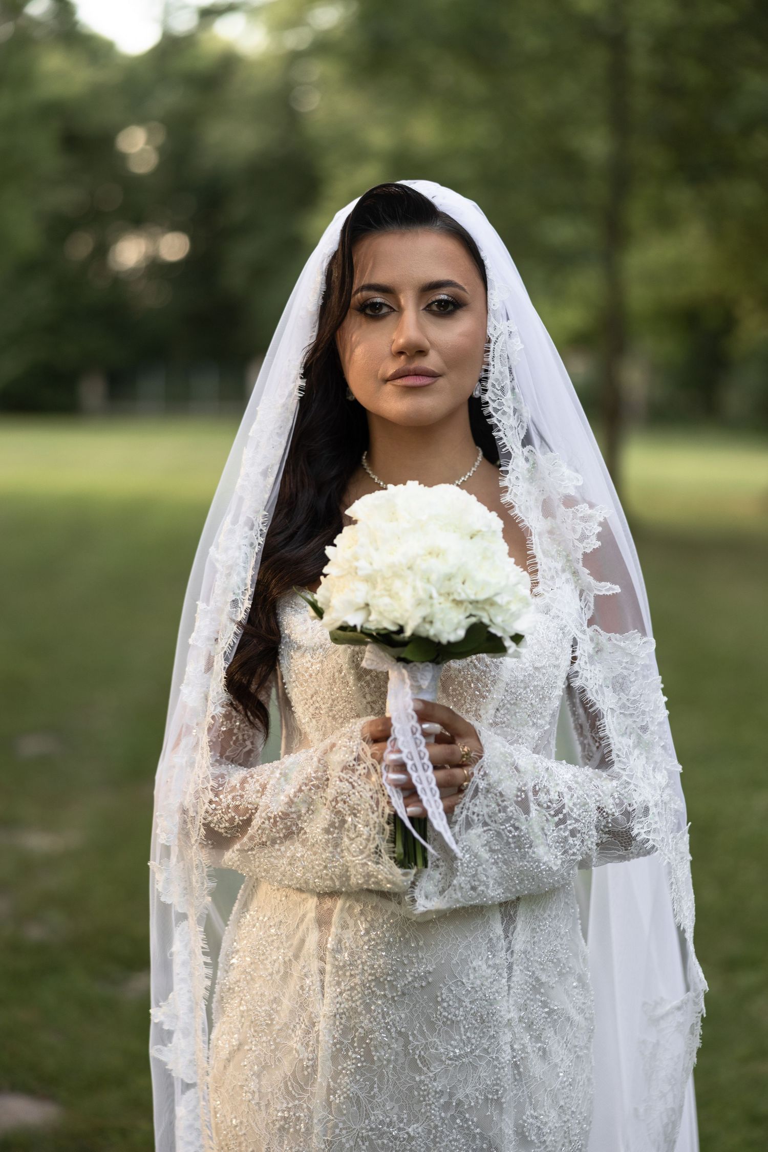 Bride in lace wedding dress and veil holds white bouquet outdoors in a green park setting.