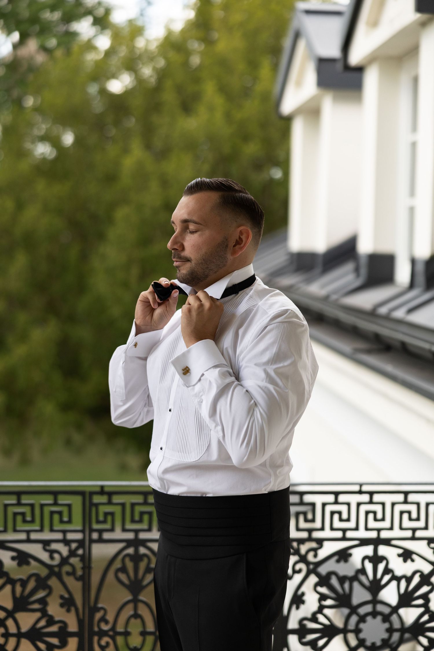 Elegant groom in white tuxedo shirt adjusting bow tie on ornate balcony with lush green trees backdrop.
