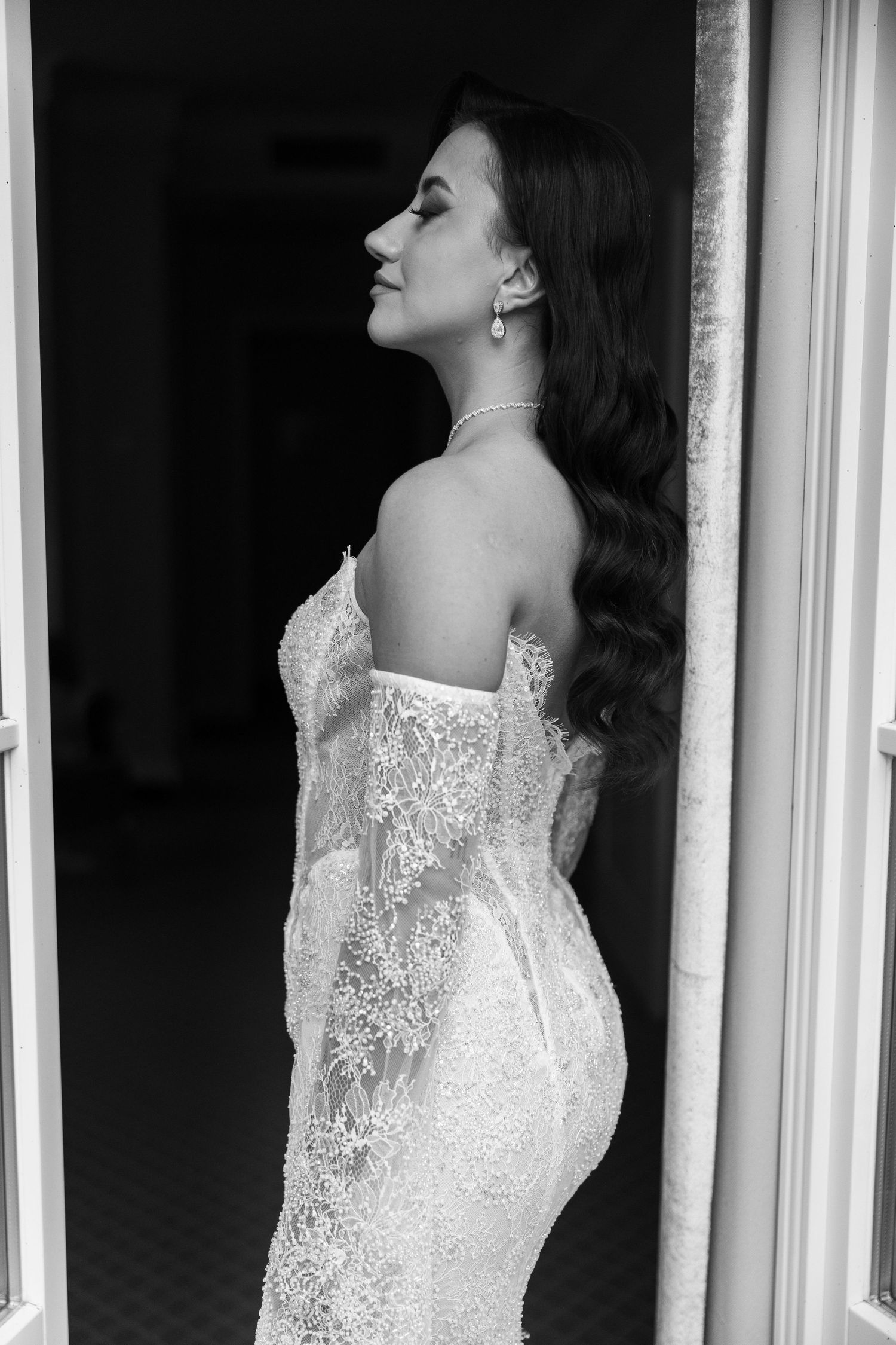 Black and white photo of a bride in a strapless lace wedding gown, standing in a doorway with elegant jewelry.