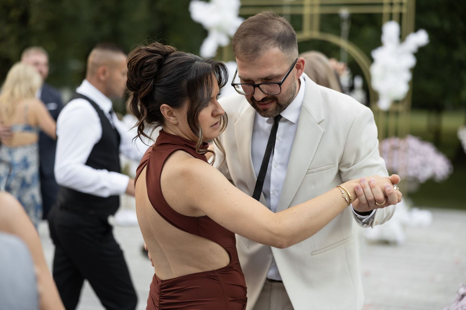 Couple dancing elegantly at an outdoor wedding, man in white suit and woman in backless burgundy dress.