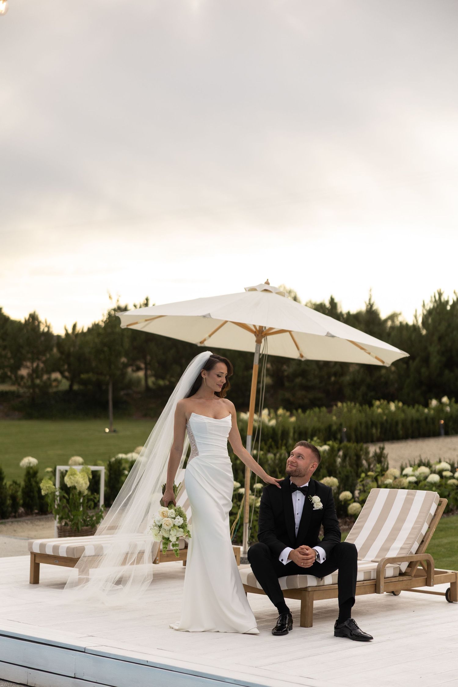 Bride in white gown and veil stands beside groom in black tuxedo seated by pool with umbrella at elegant outdoor venue.