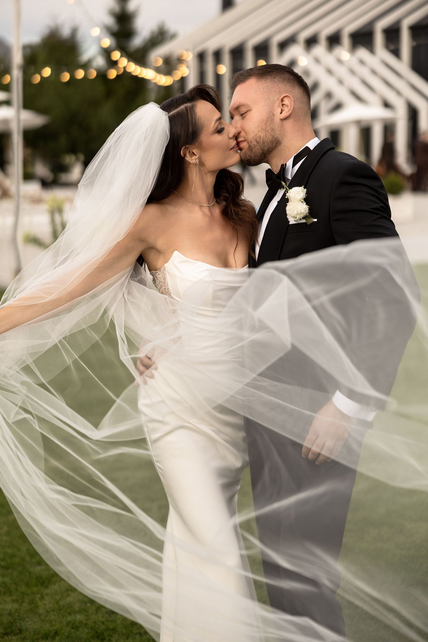 Bride and groom share a romantic kiss outdoors, her flowing white veil billowing around them at their elegant wedding venue.