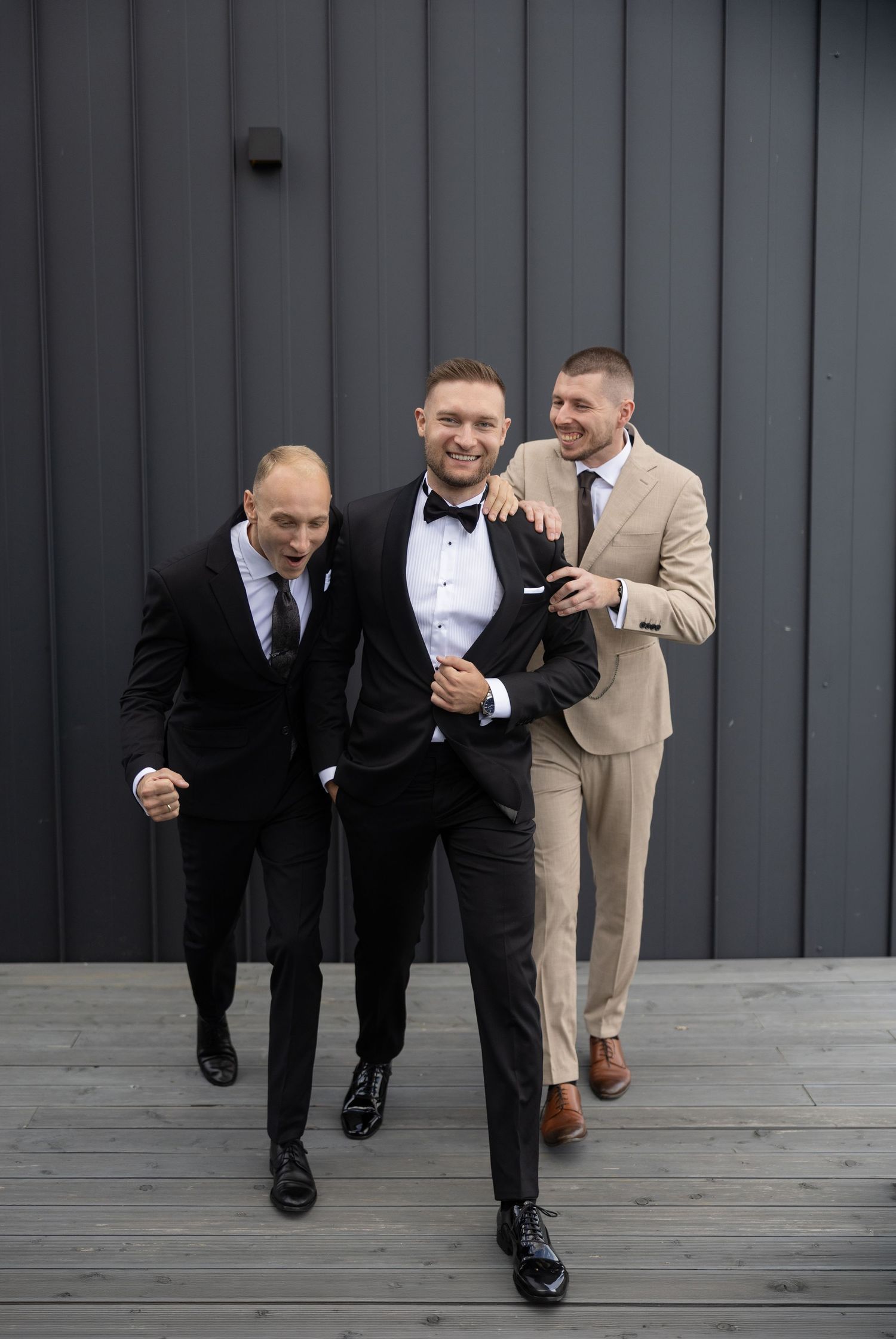 Three men in suits pose playfully outside, with the groom in a black tuxedo flanked by smiling groomsmen.
