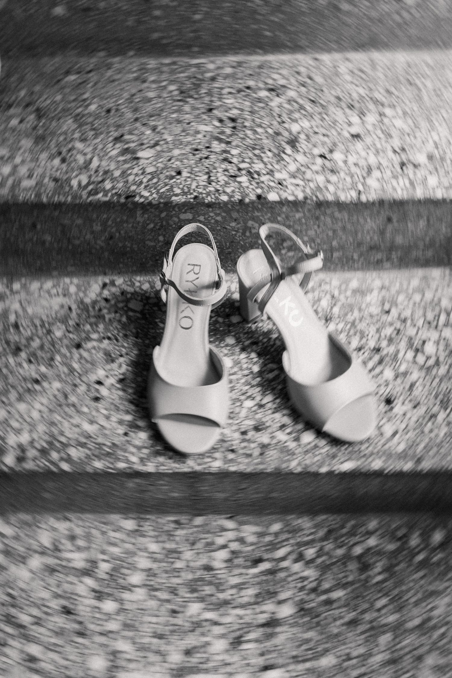 A pair of light-colored women's sandals with ankle straps photographed in black and white on a speckled surface.