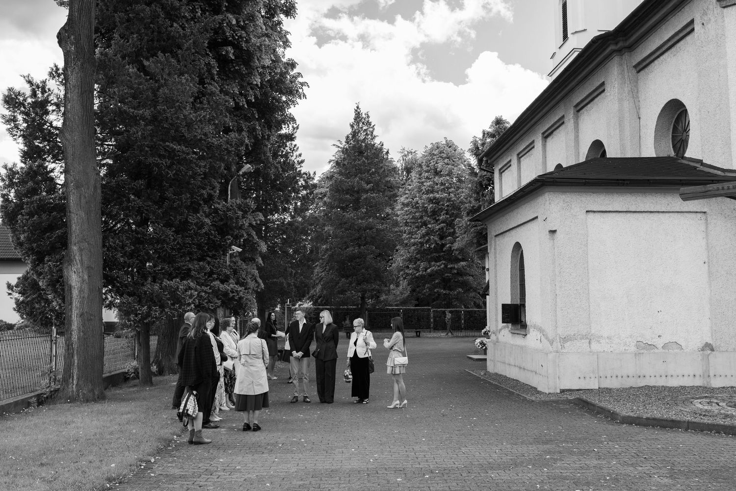 A group of people walking along a tree-lined path next to a white church building in a vintage black and white photo.