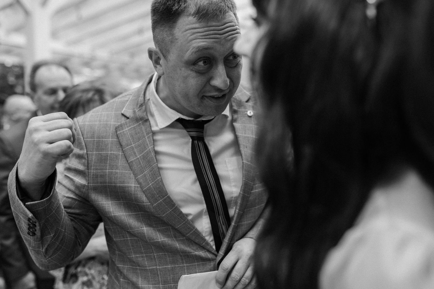 A black and white photograph shows a person in a plaid suit jacket gesturing while engaged in conversation.