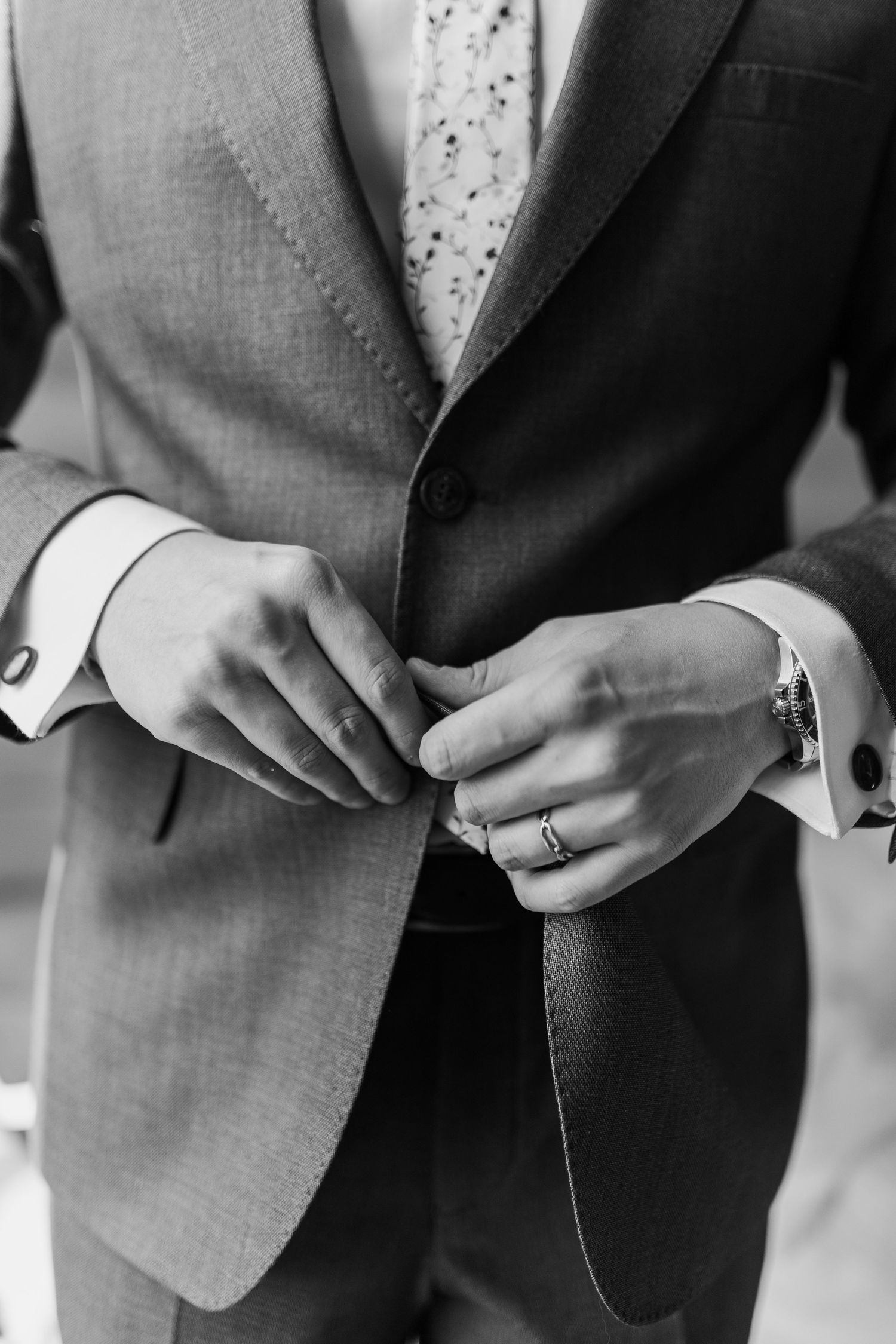 Black and white close-up of groom's hands buttoning suit jacket, wearing wedding ring and cufflinks.