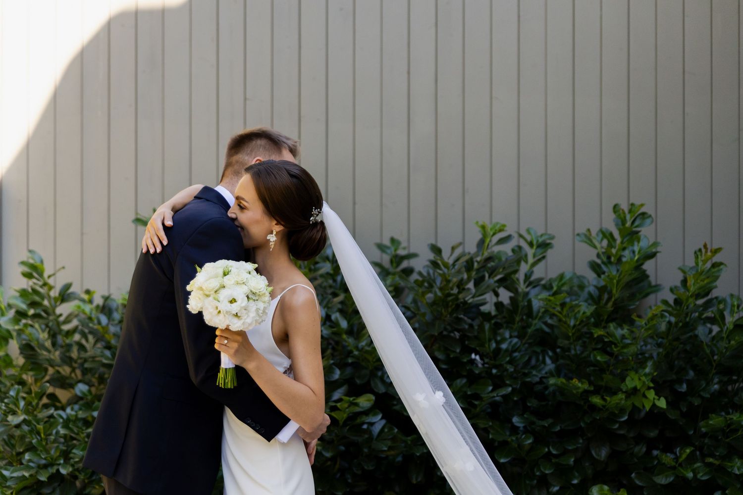 A newlywed couple embraces outdoors with a white bouquet against a gray wall and green shrubs.