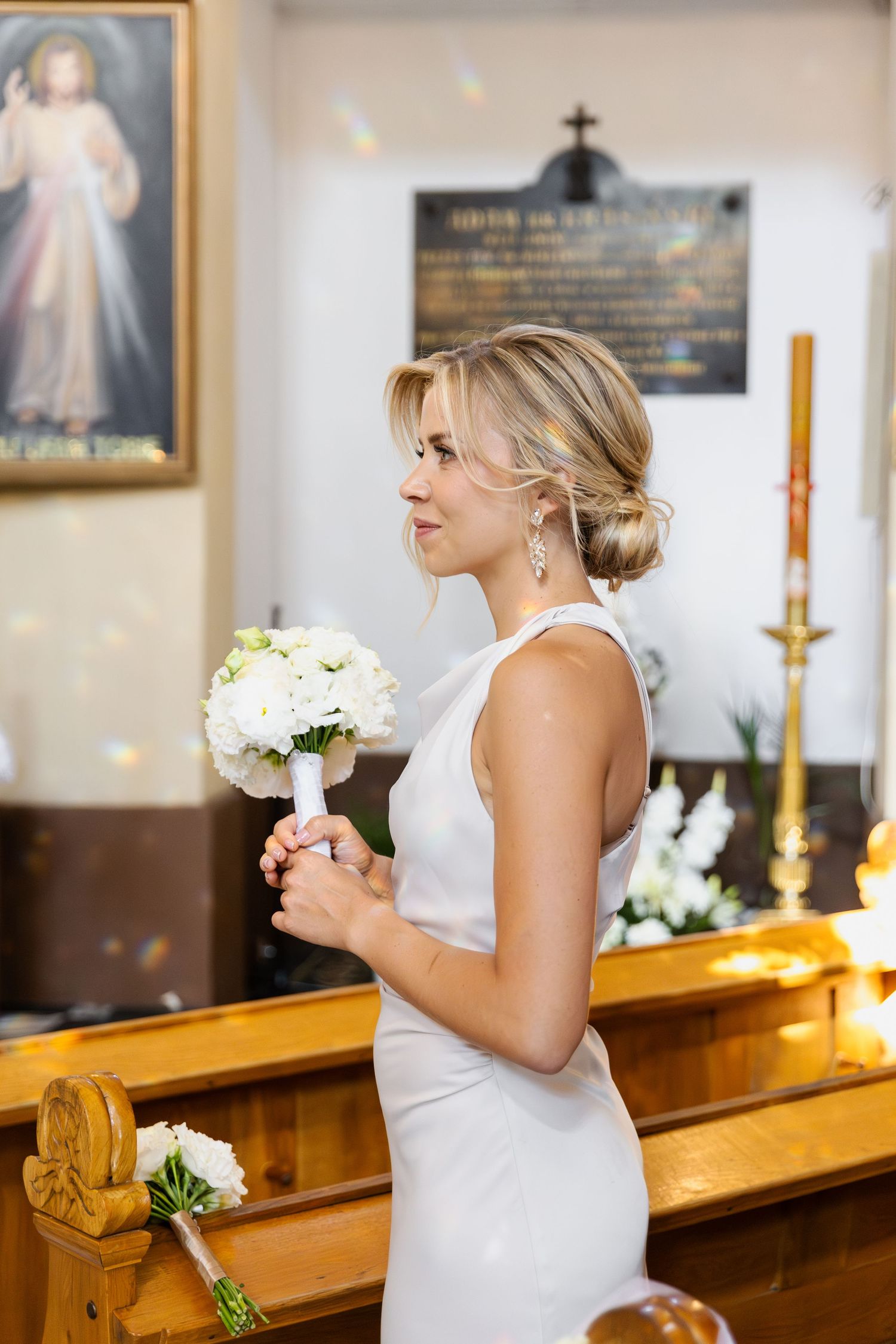 A bride in an elegant white sleeveless dress holds a bouquet of white flowers while standing in a church sanctuary.