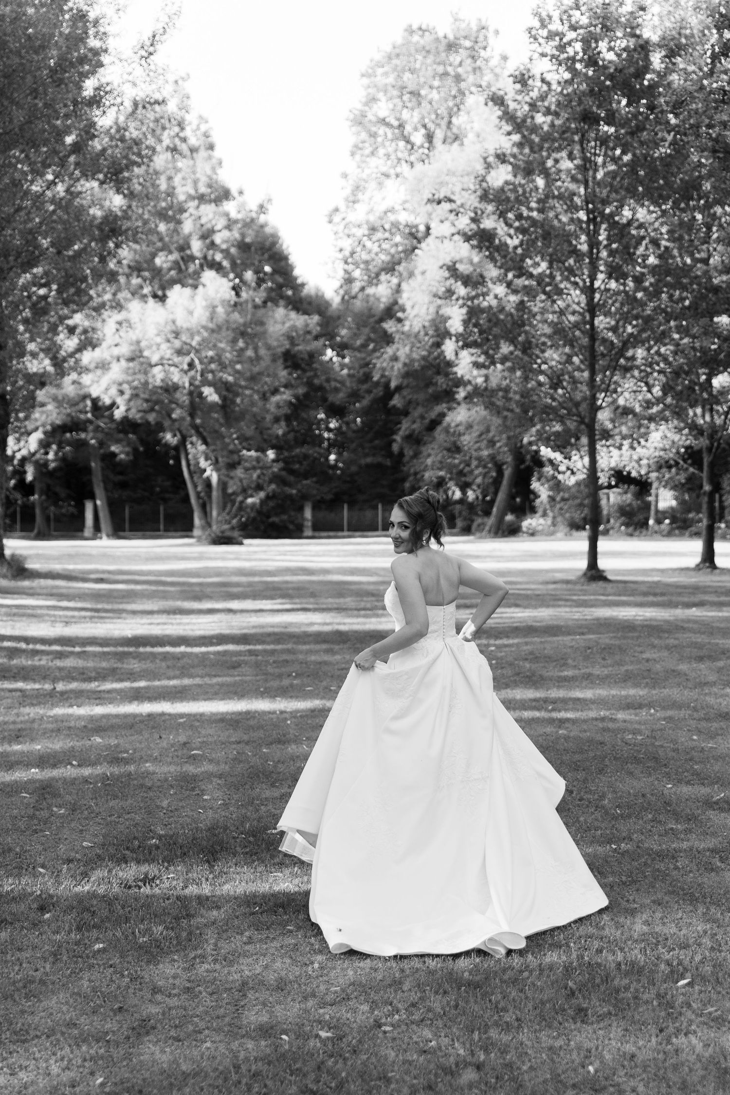 A person in an elegant white wedding gown twirls on a grassy lawn surrounded by trees in a black and white photograph.