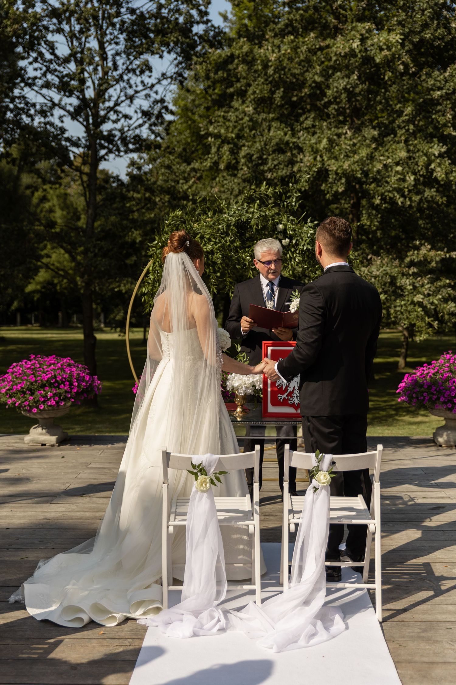 A wedding ceremony taking place outdoors with white chairs and vibrant pink flowers in the background.