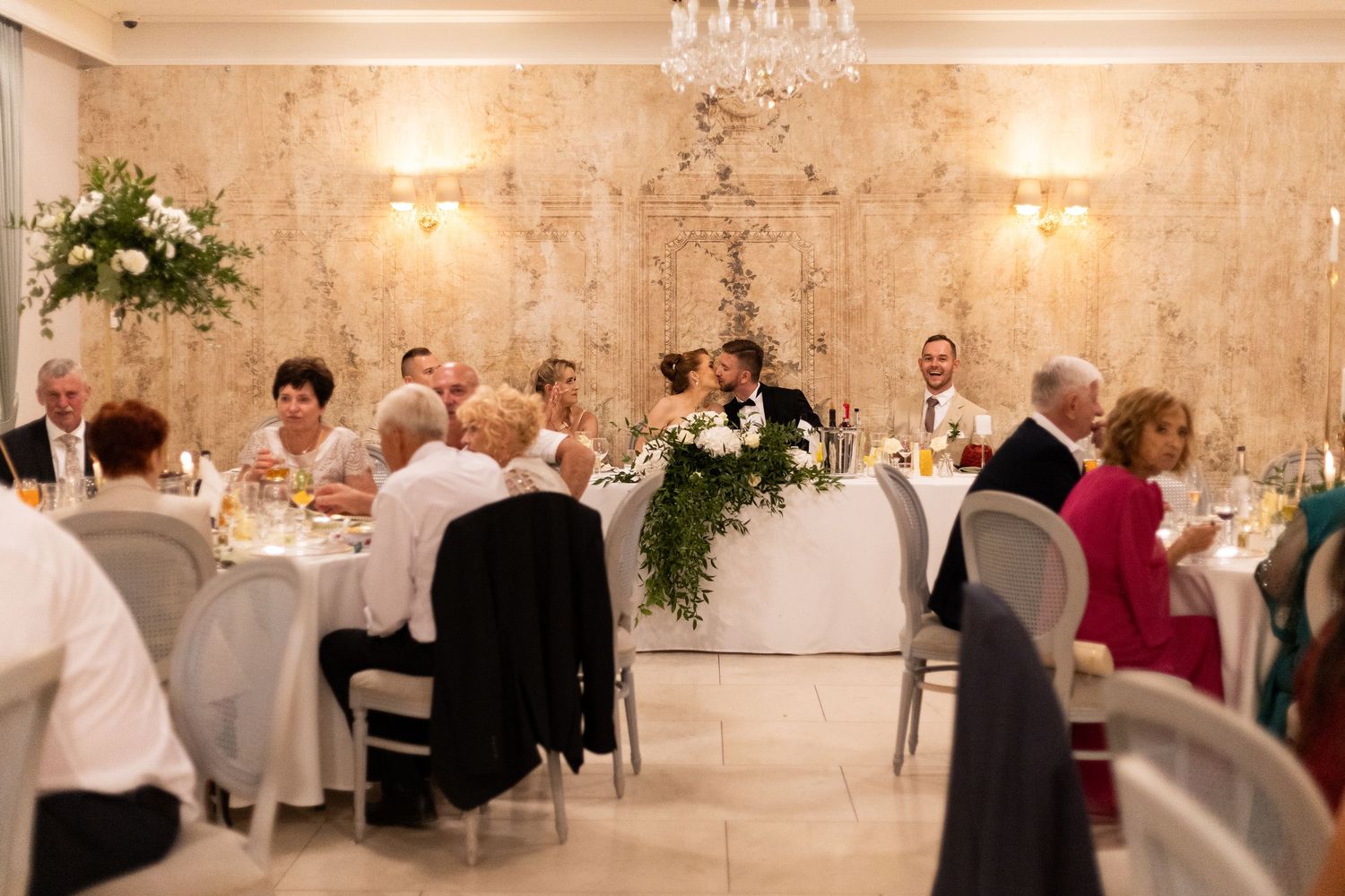 A panoramic view of an elegant wedding reception venue with crystal chandeliers, white decor and guests seated at tables.