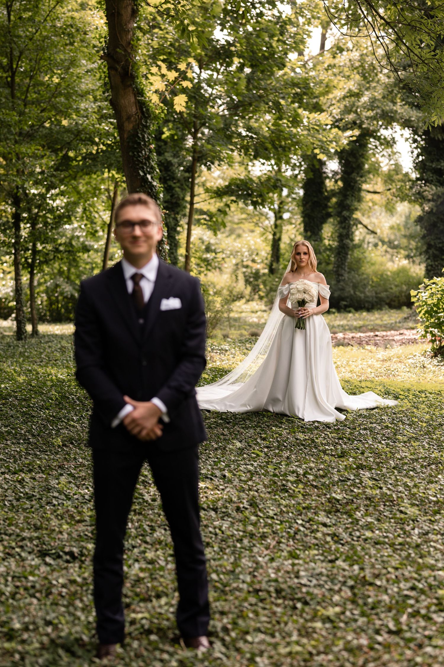 Bride and groom stand apart in a wooded area covered with fallen leaves during their first look wedding photos.