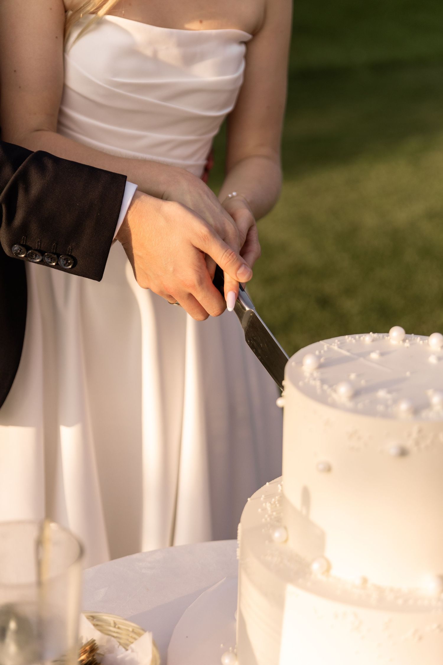 People in white dresses cutting into an elegant white wedding cake at an outdoor reception.