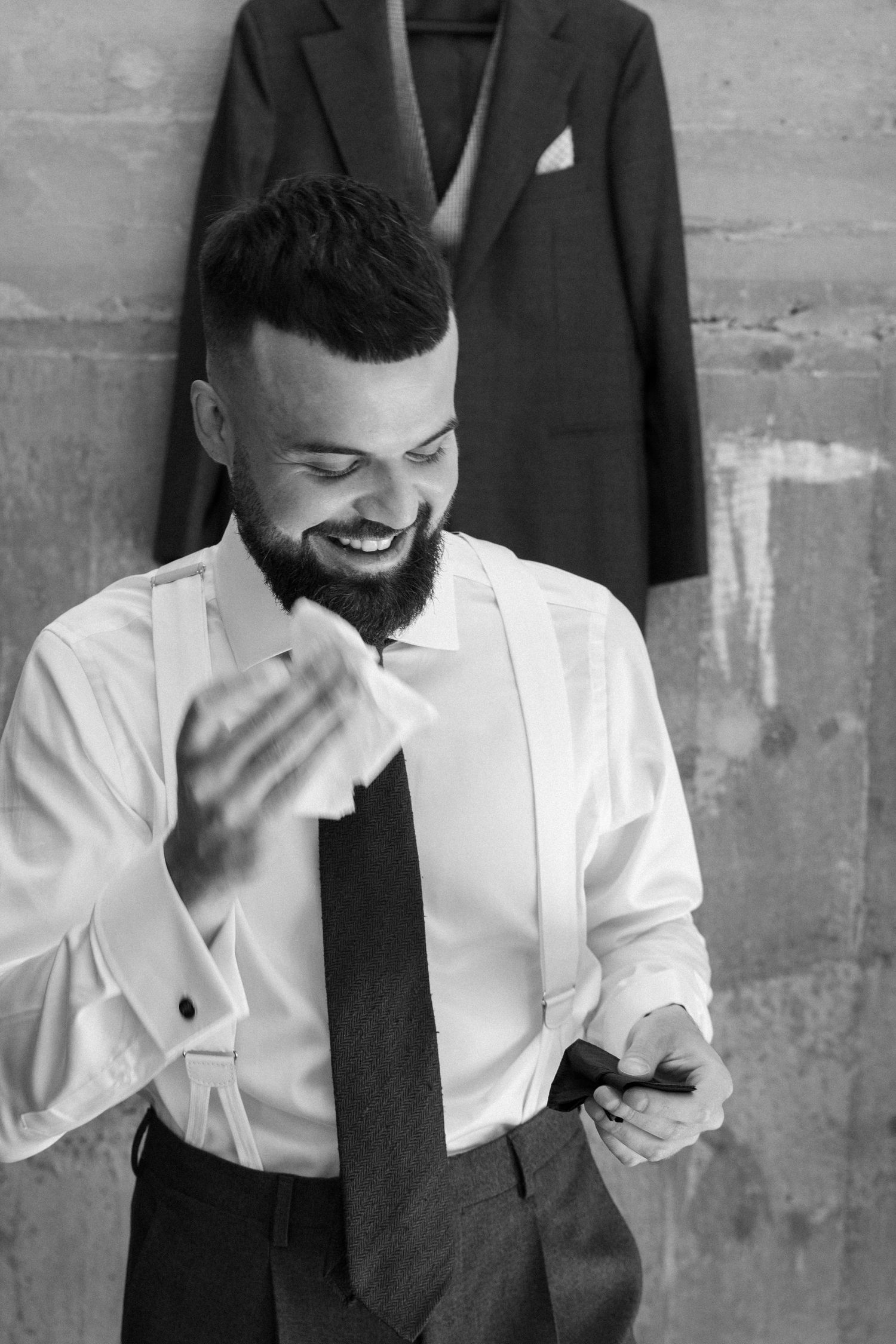 A smiling groom in a white dress shirt and black tie looks down at his phone with a suit jacket hanging in the background.