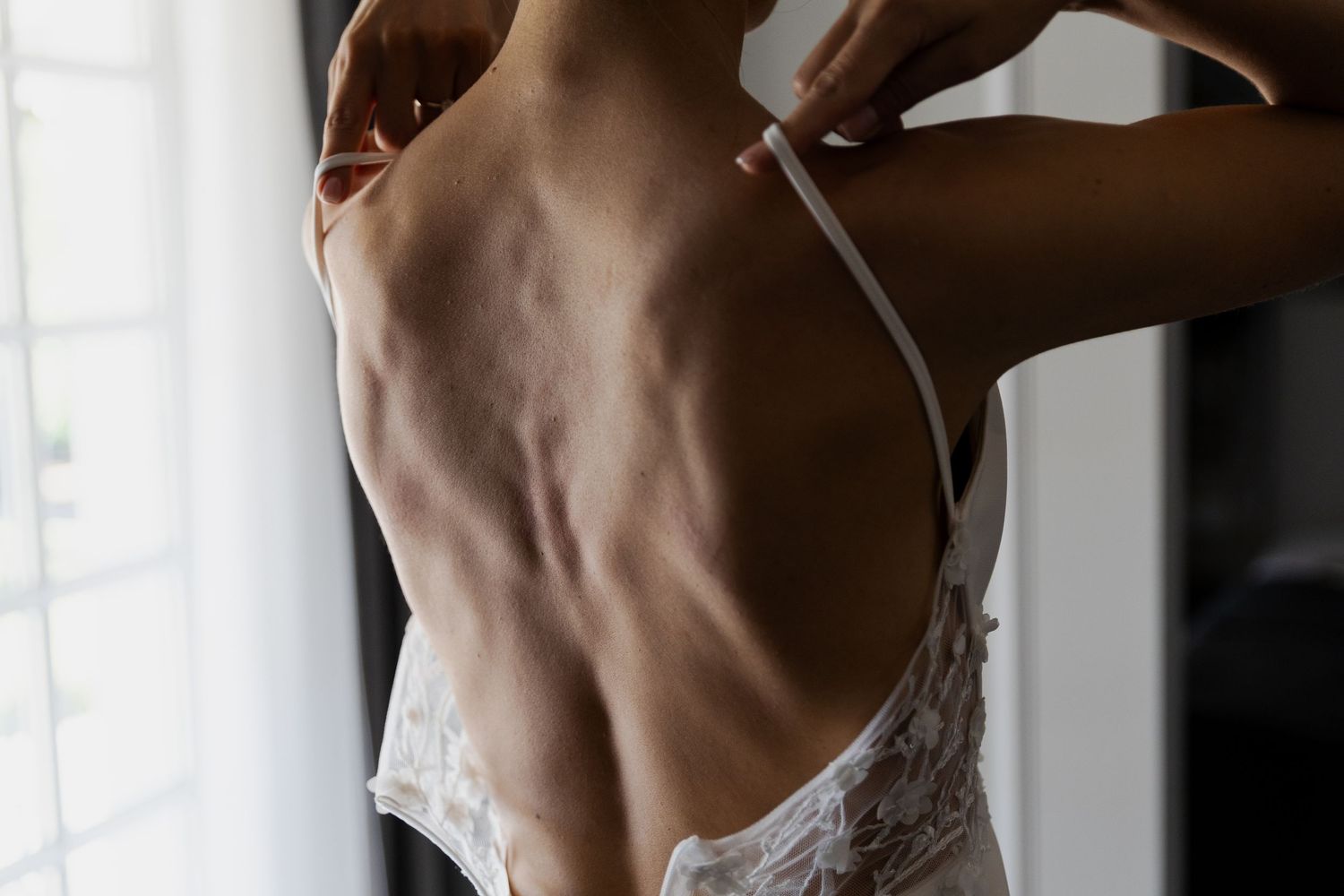 Artistic close up of a person's back wearing delicate white lace lingerie near a window in soft natural light.