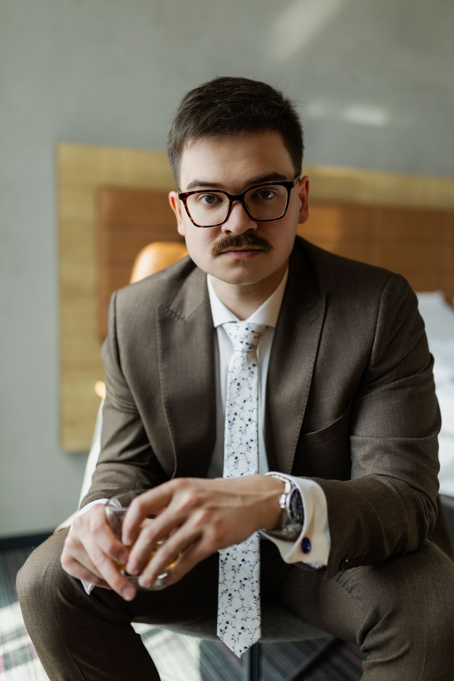 Professional portrait of someone in a brown suit and patterned tie sitting in an office setting with glasses and mustache.