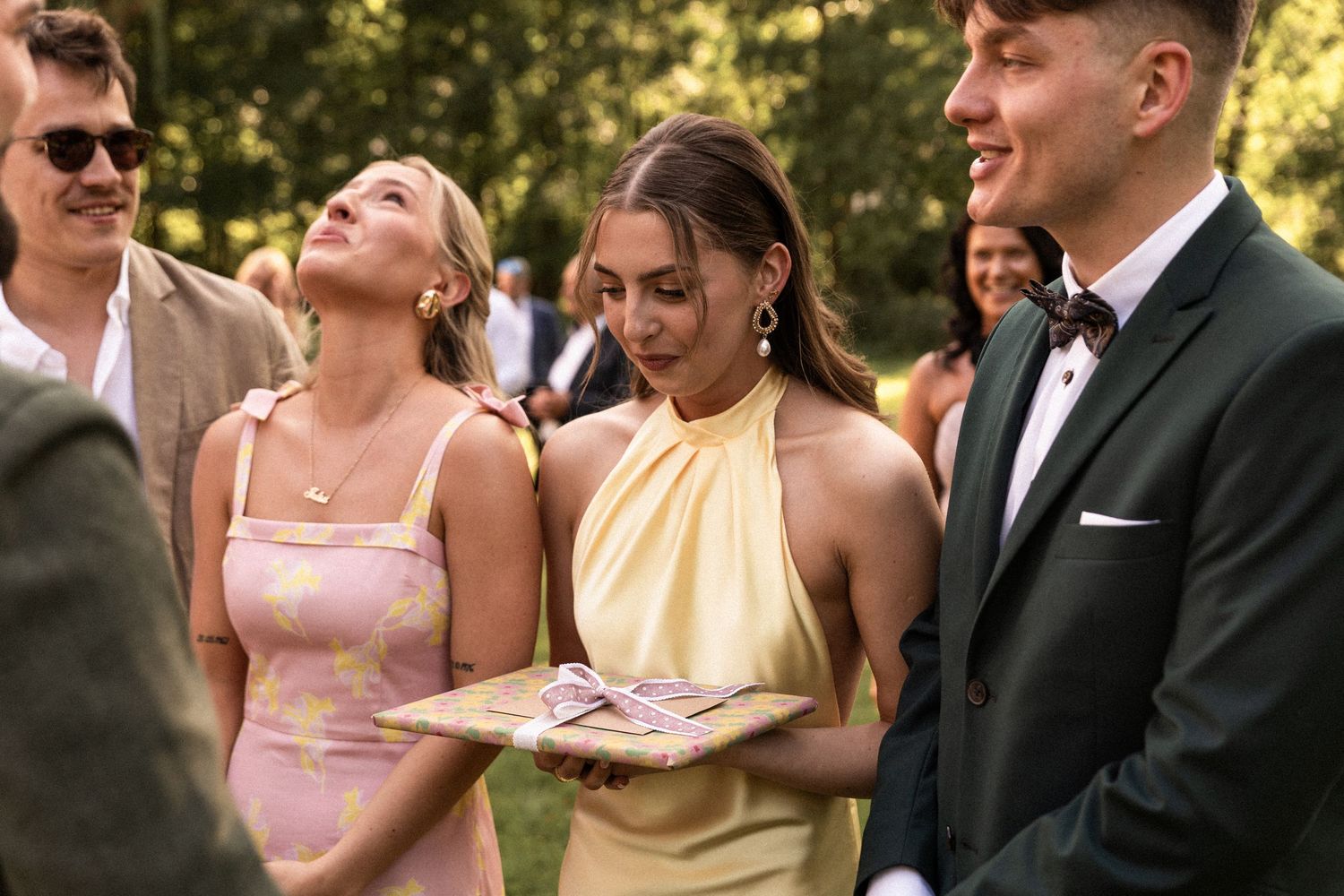 Smiling wedding party members in formal attire gather outdoors during a sunny summer celebration.
