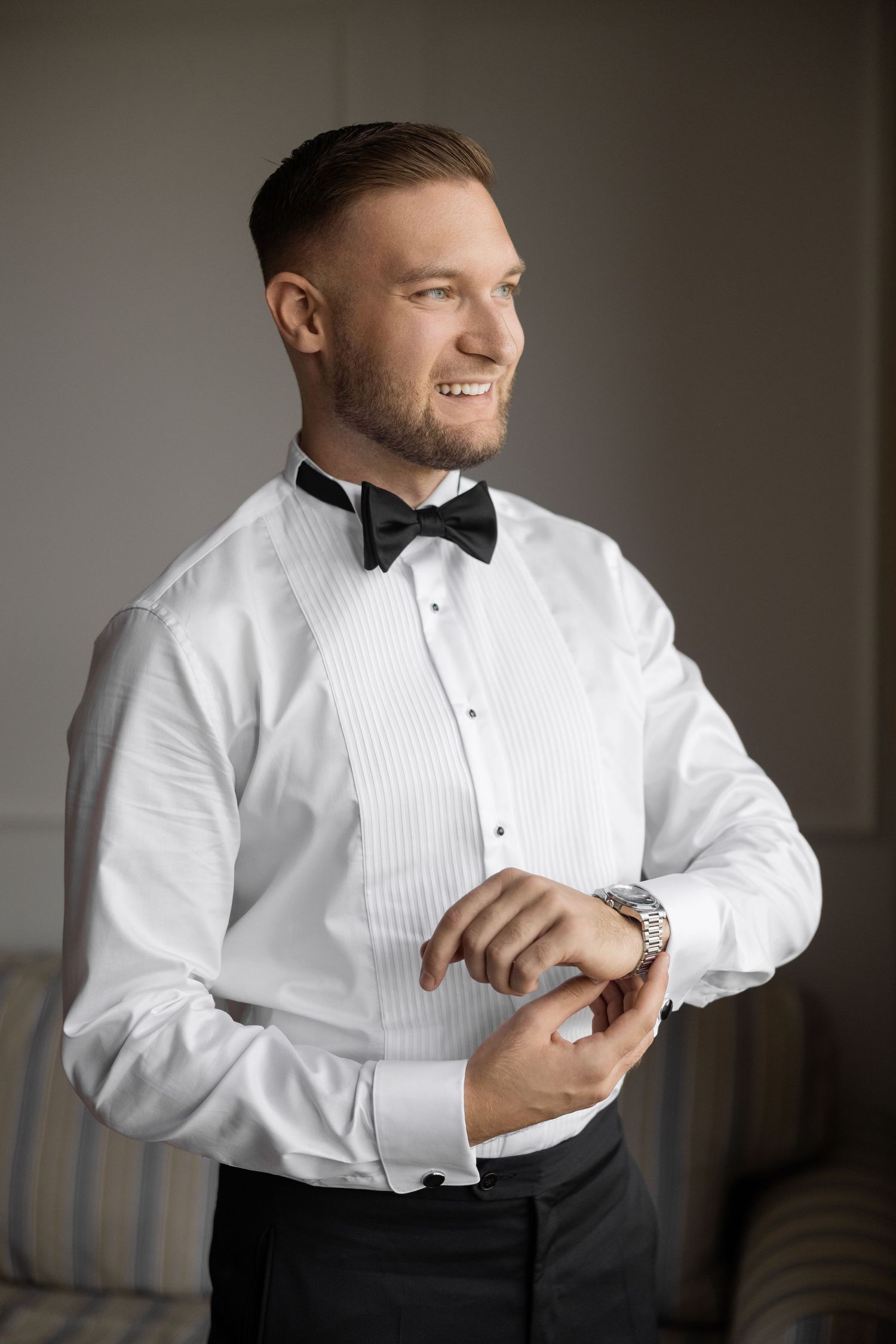 Smiling groom in white tuxedo shirt and black bow tie adjusting his watch cufflinks before wedding ceremony.