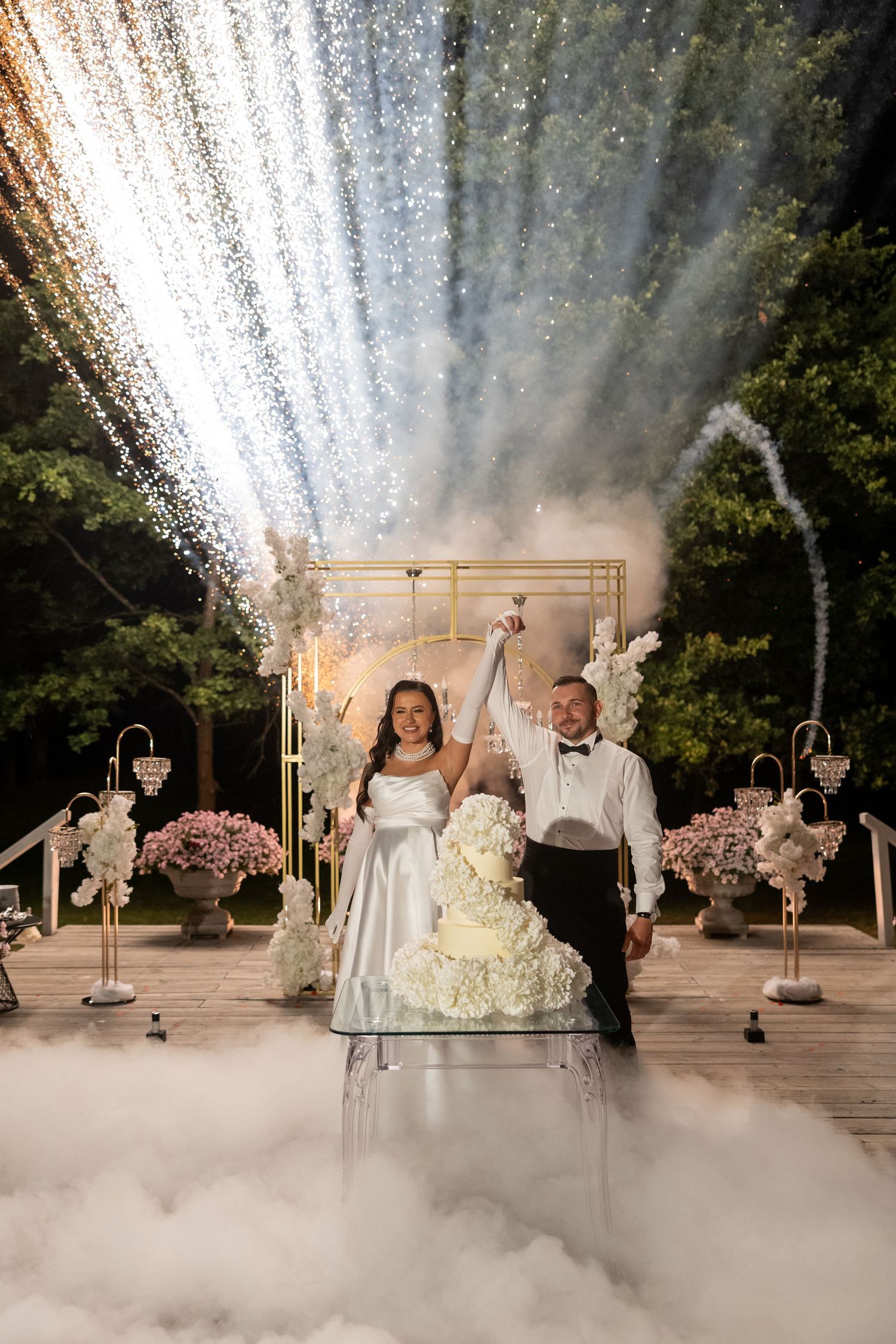 Bride and groom cutting wedding cake outdoors at night with sparkling fireworks display and misty fog effects.