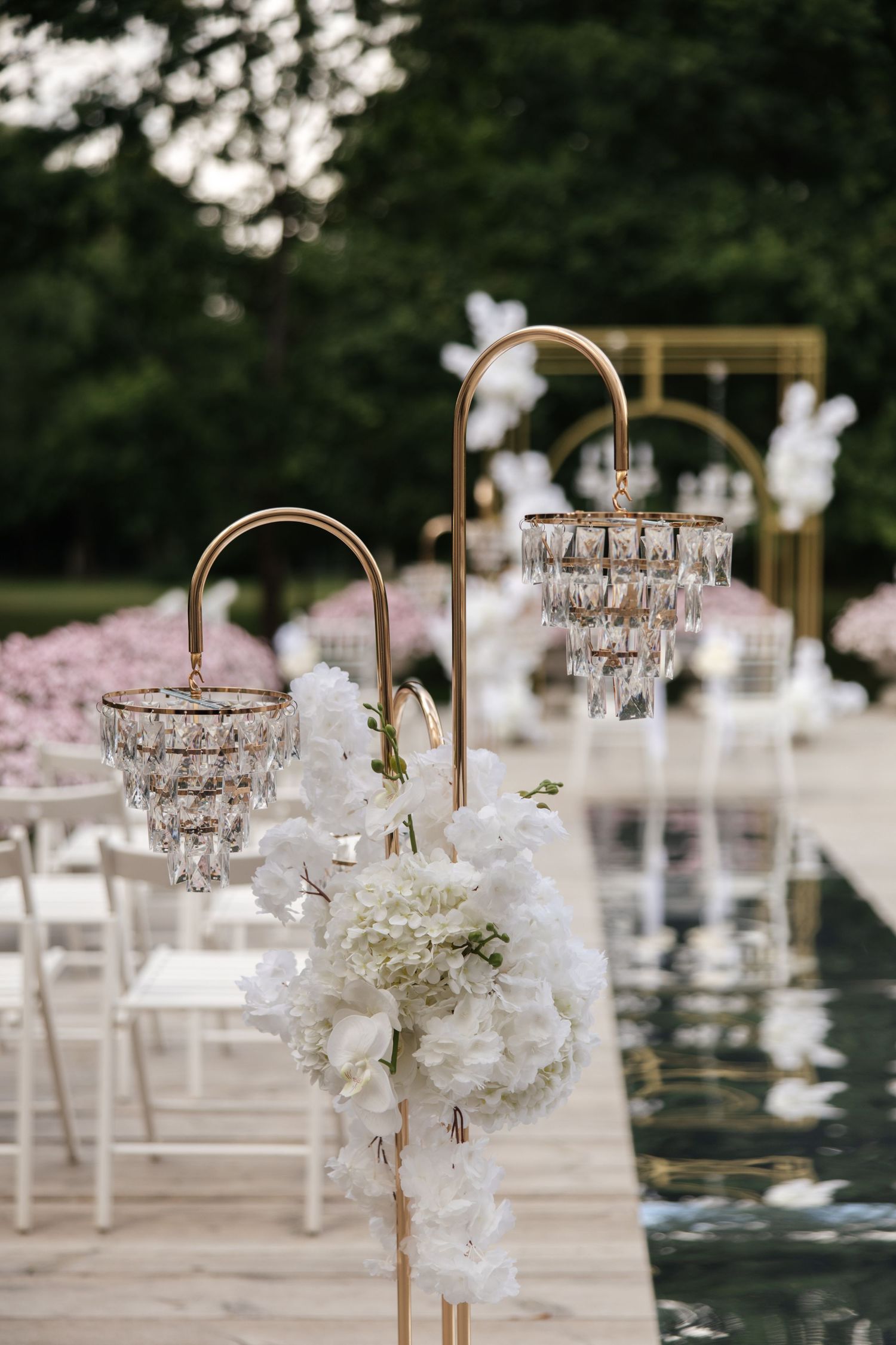 Elegant outdoor wedding aisle with gold crystal chandeliers, white hydrangeas, and chairs beside a reflective pool.