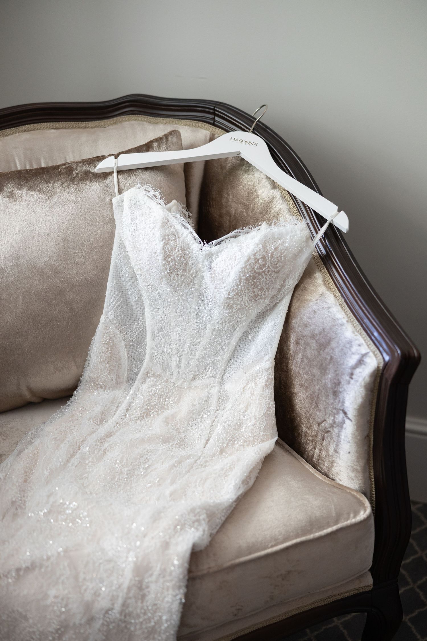 Elegant white lace strapless wedding dress on a hanger draped over a vintage cream velvet sofa.