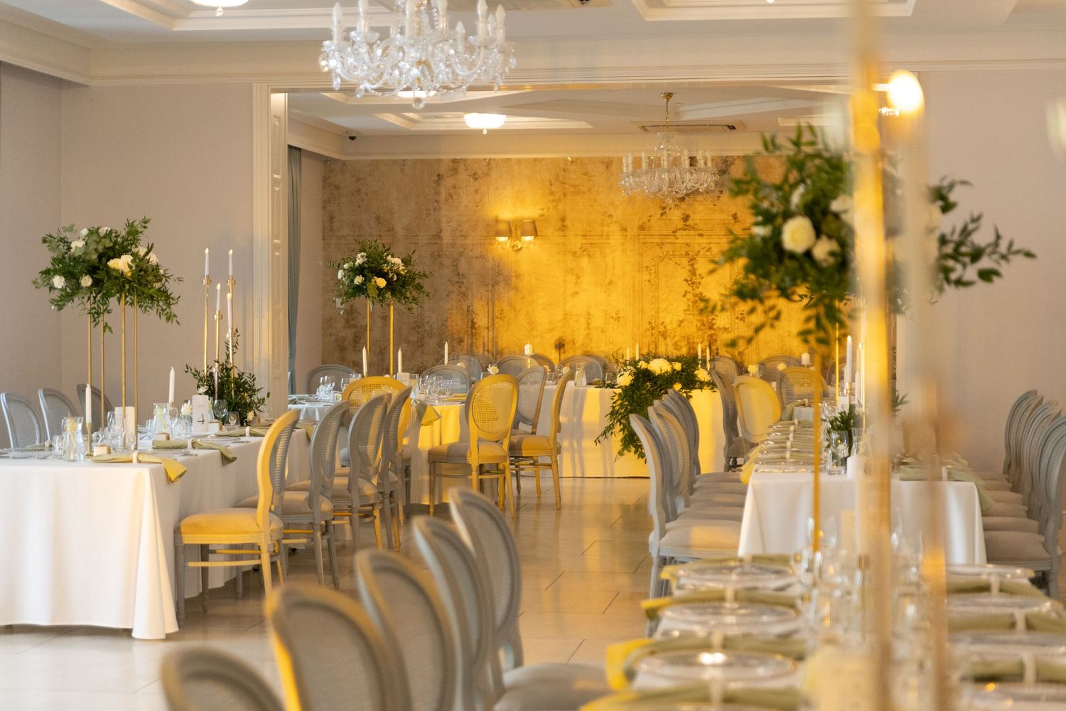 An elegant wedding reception venue with crystal chandeliers, white tablecloths, and floral centerpieces in a bright room.