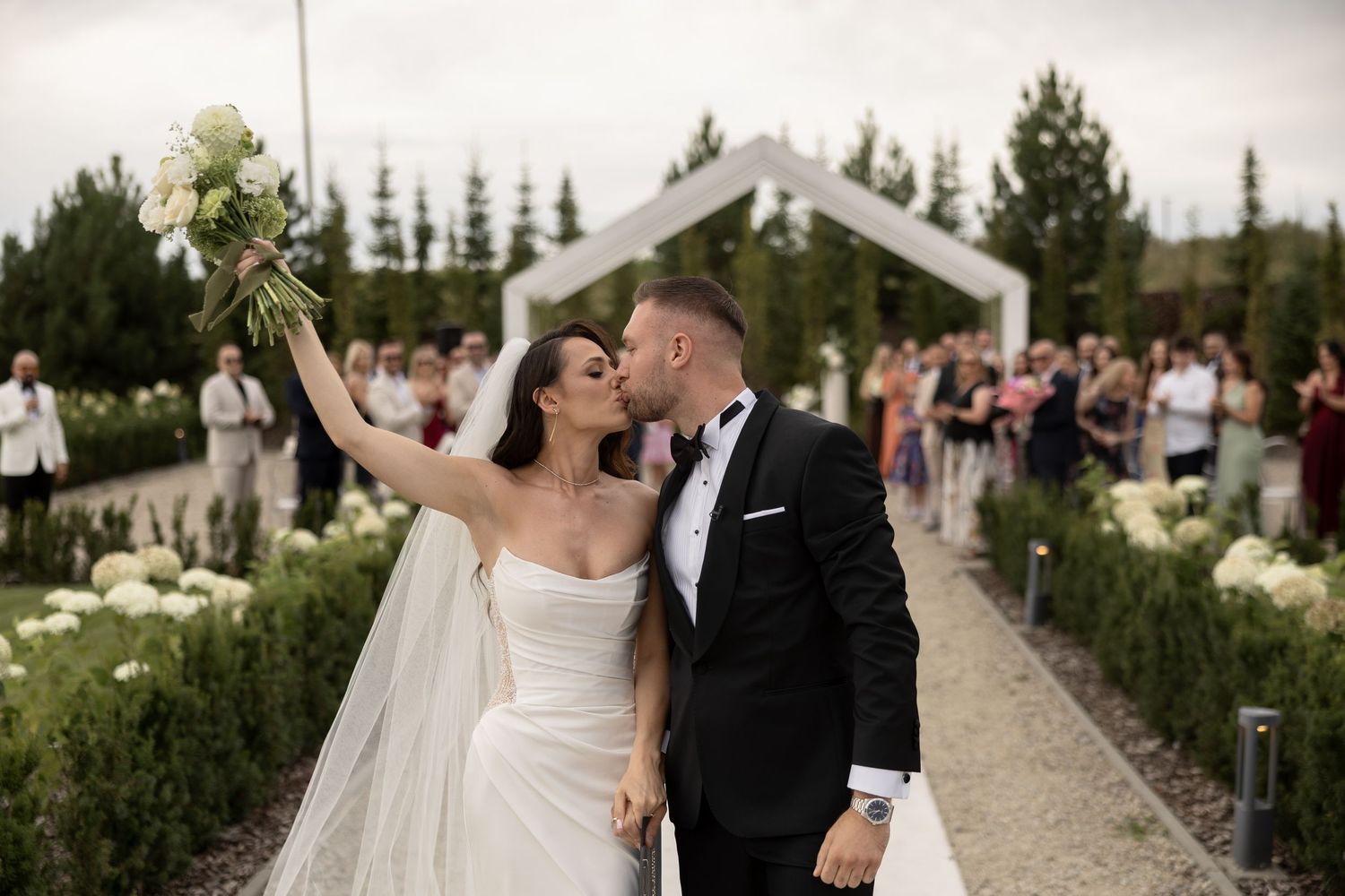 Newlyweds kiss on aisle as bride raises bouquet with guests in background.