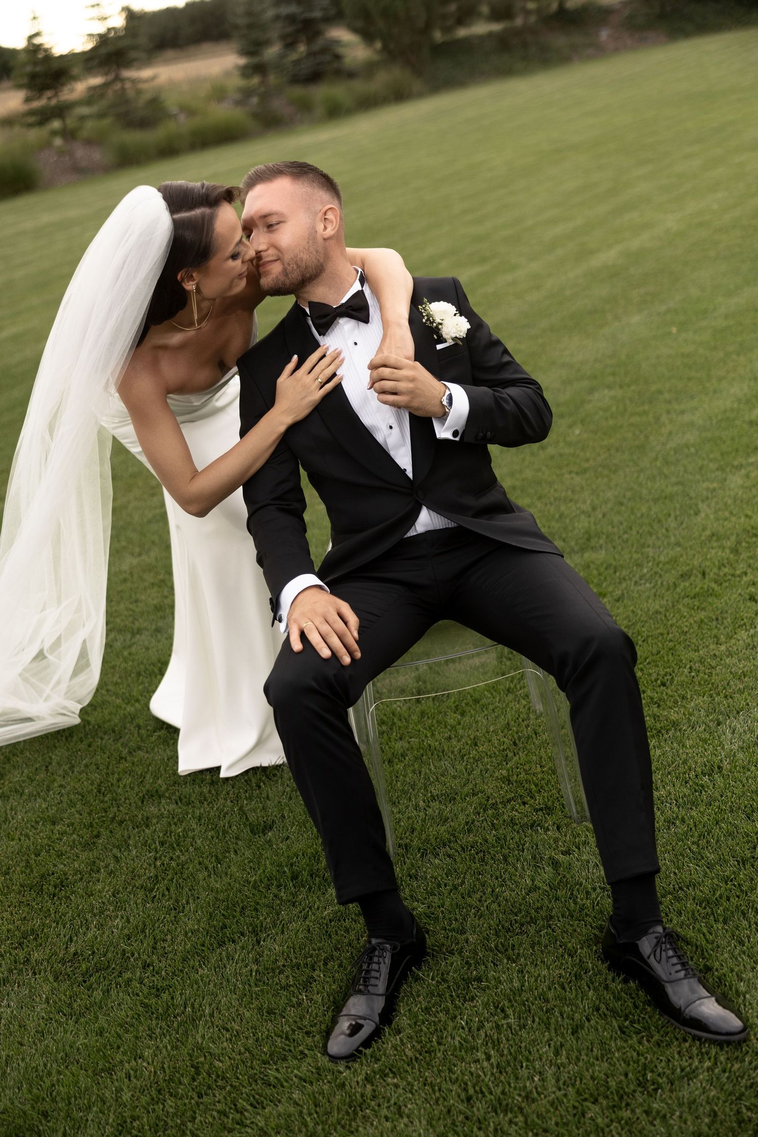 Bride in white gown kisses groom in black tuxedo sitting on chair on green lawn at outdoor wedding.