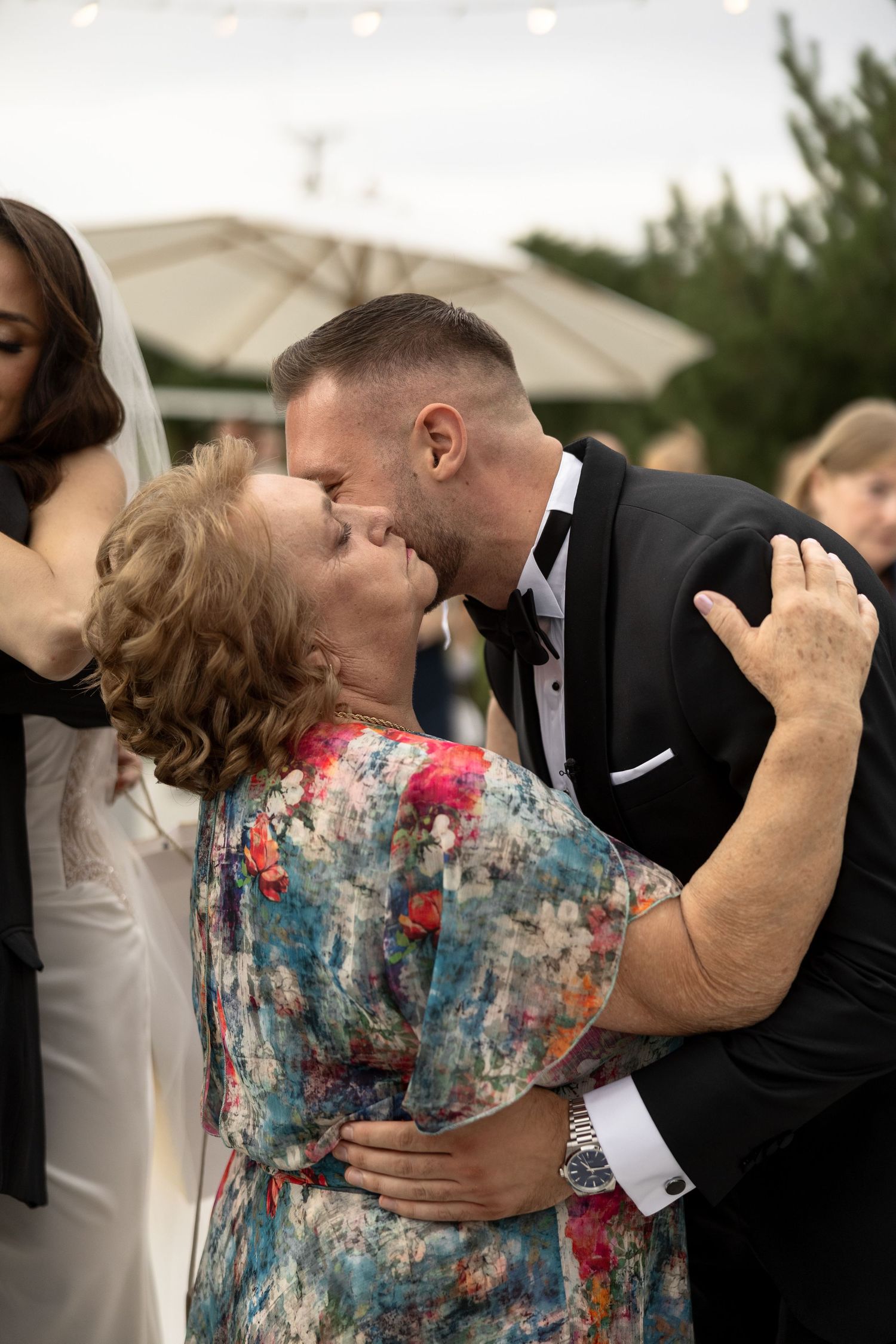 Groom in black tuxedo shares a tender kiss with older woman in floral dress at outdoor wedding reception.