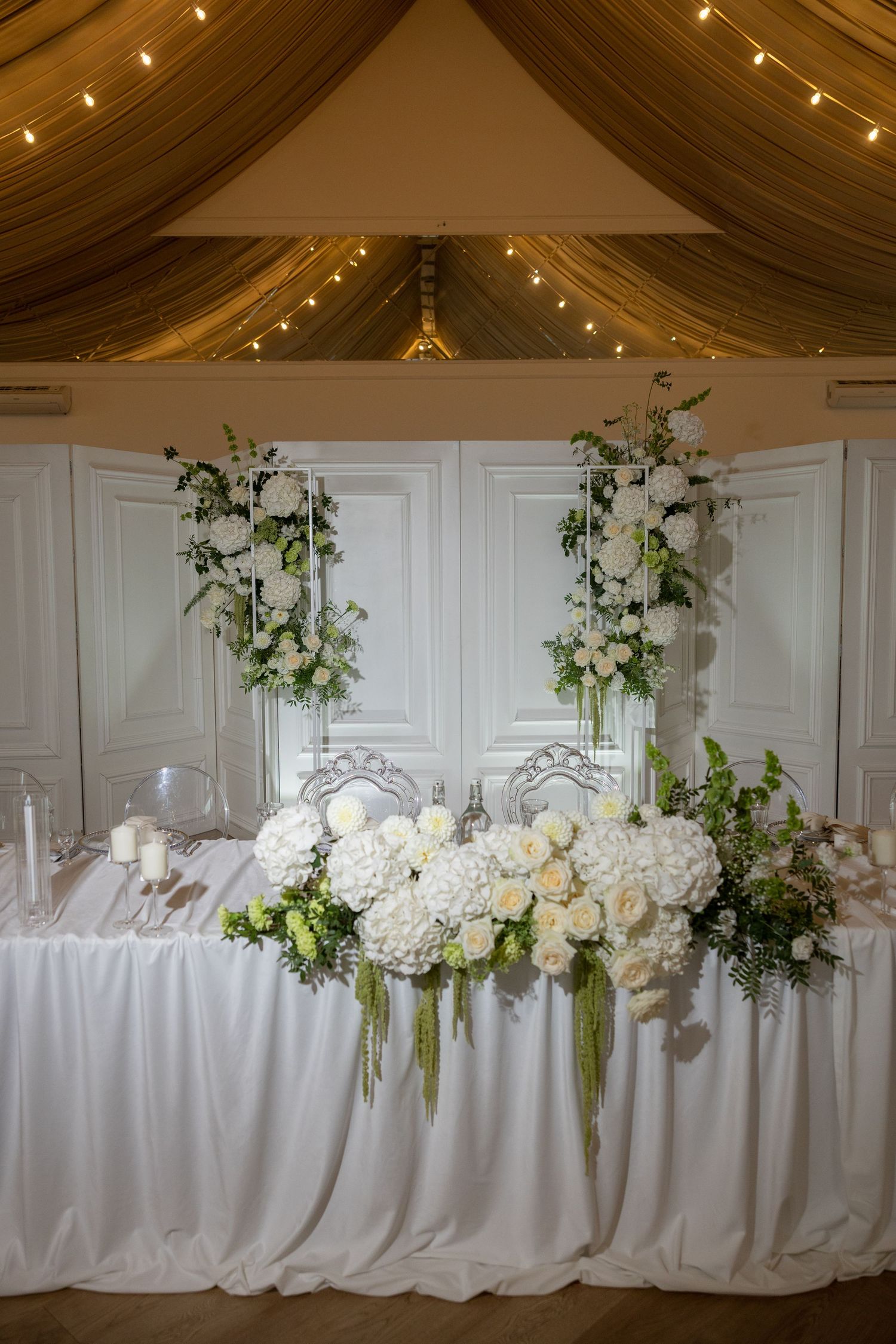 Elegant wedding sweetheart table with white floral arrangements, candles, and draped fabric ceiling with fairy lights.
