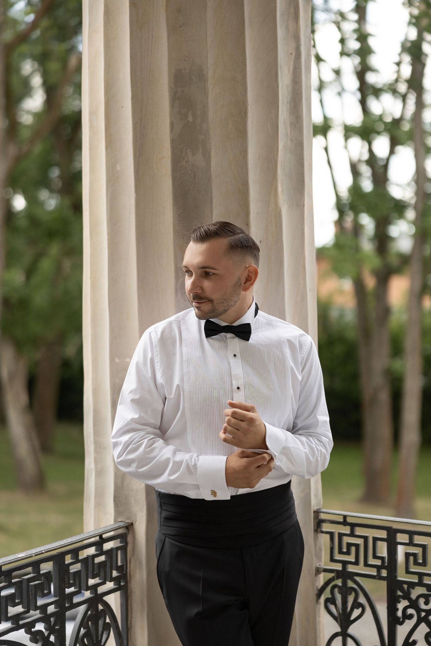 Elegant groom in white tuxedo shirt and black bow tie adjusting cufflinks on a decorative balcony.