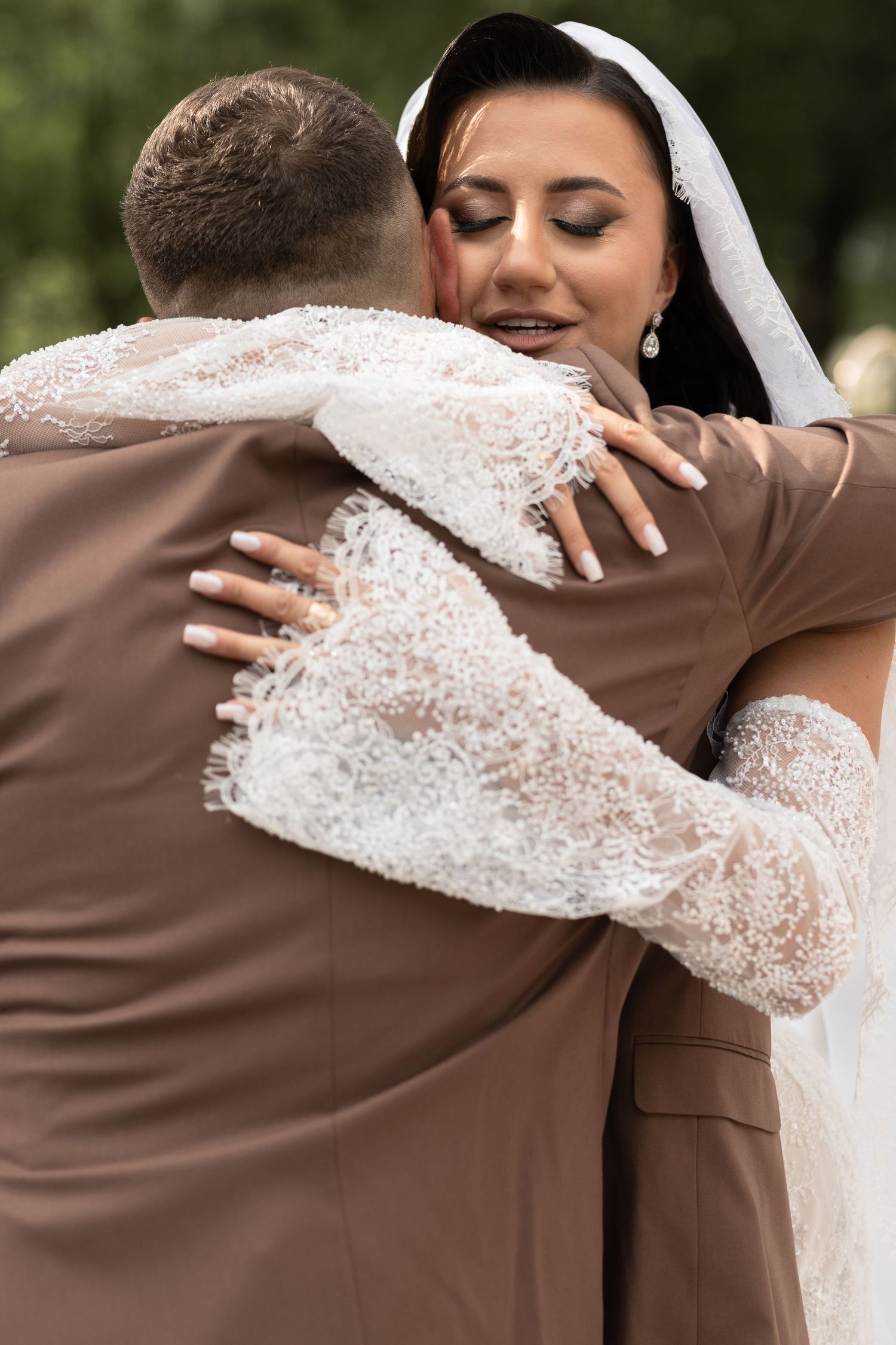 Bride in white lace dress and veil embraces groom in brown suit outdoors during wedding ceremony.