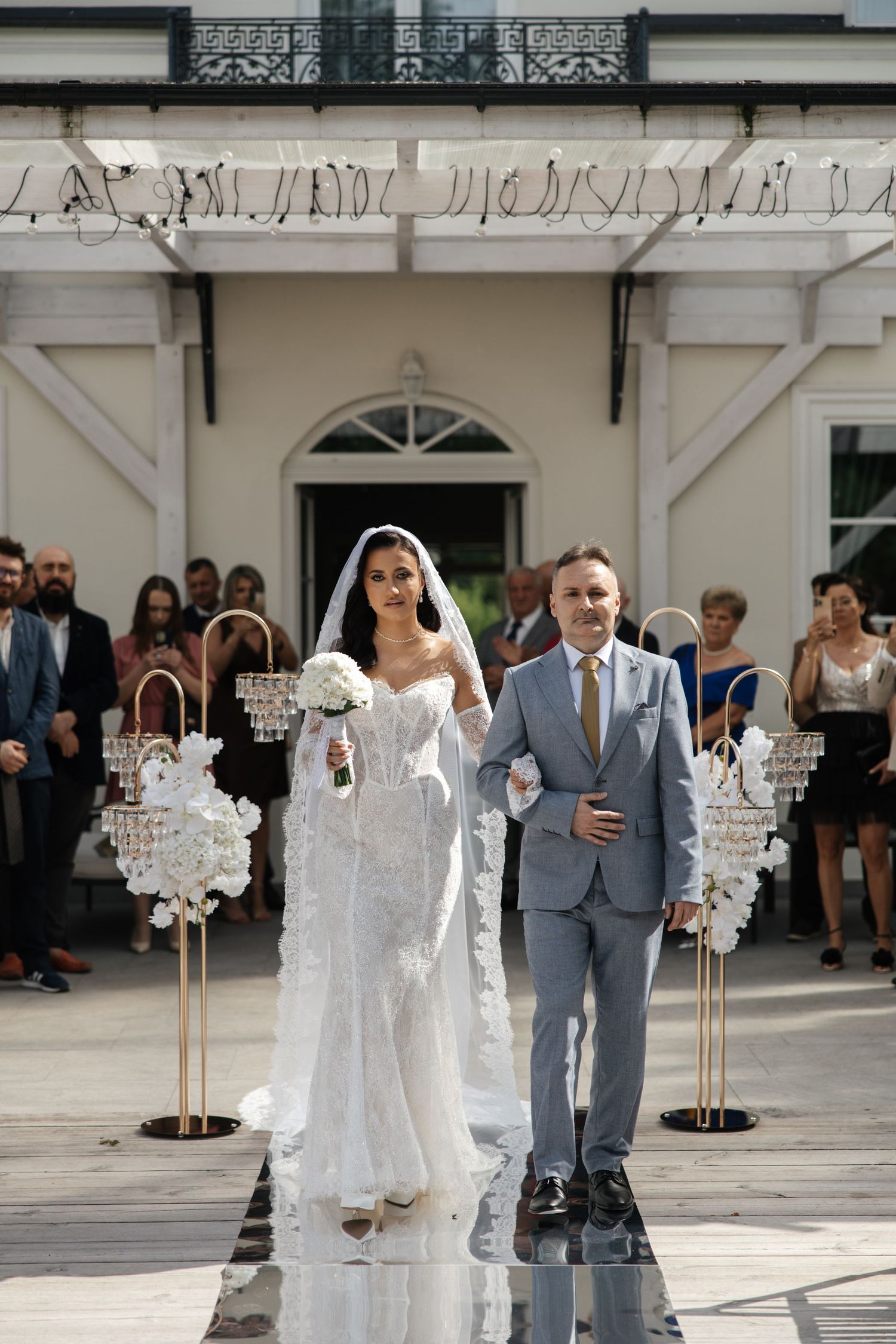 Bride in white lace gown and veil walks down aisle with escort at elegant outdoor wedding ceremony.