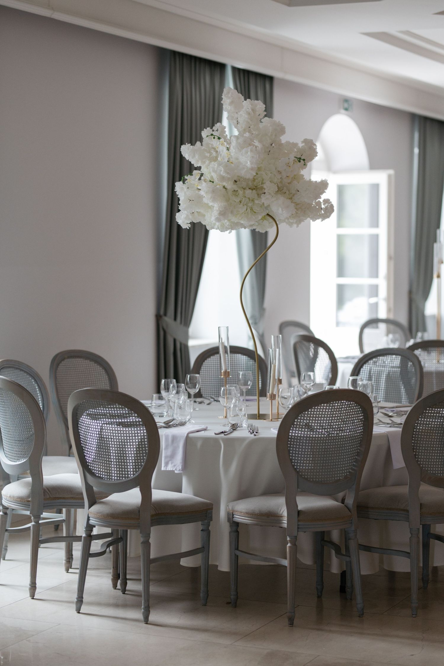 Elegant wedding reception table with white floral centerpiece, gray Louis chairs, and glassware in a bright venue.