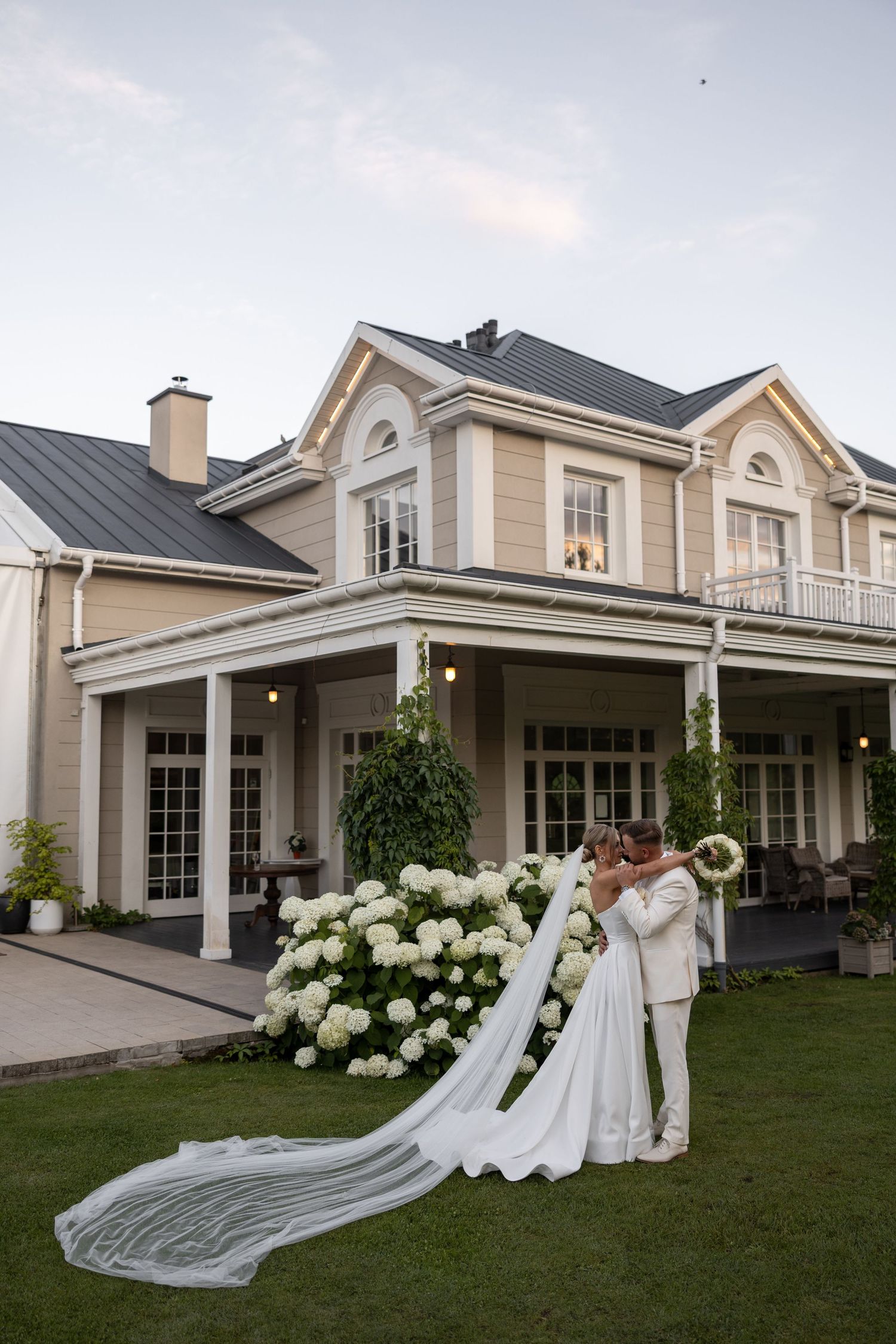 Bride and groom kissing in front of elegant manor house with white hydrangeas and long bridal veil trailing on lawn.