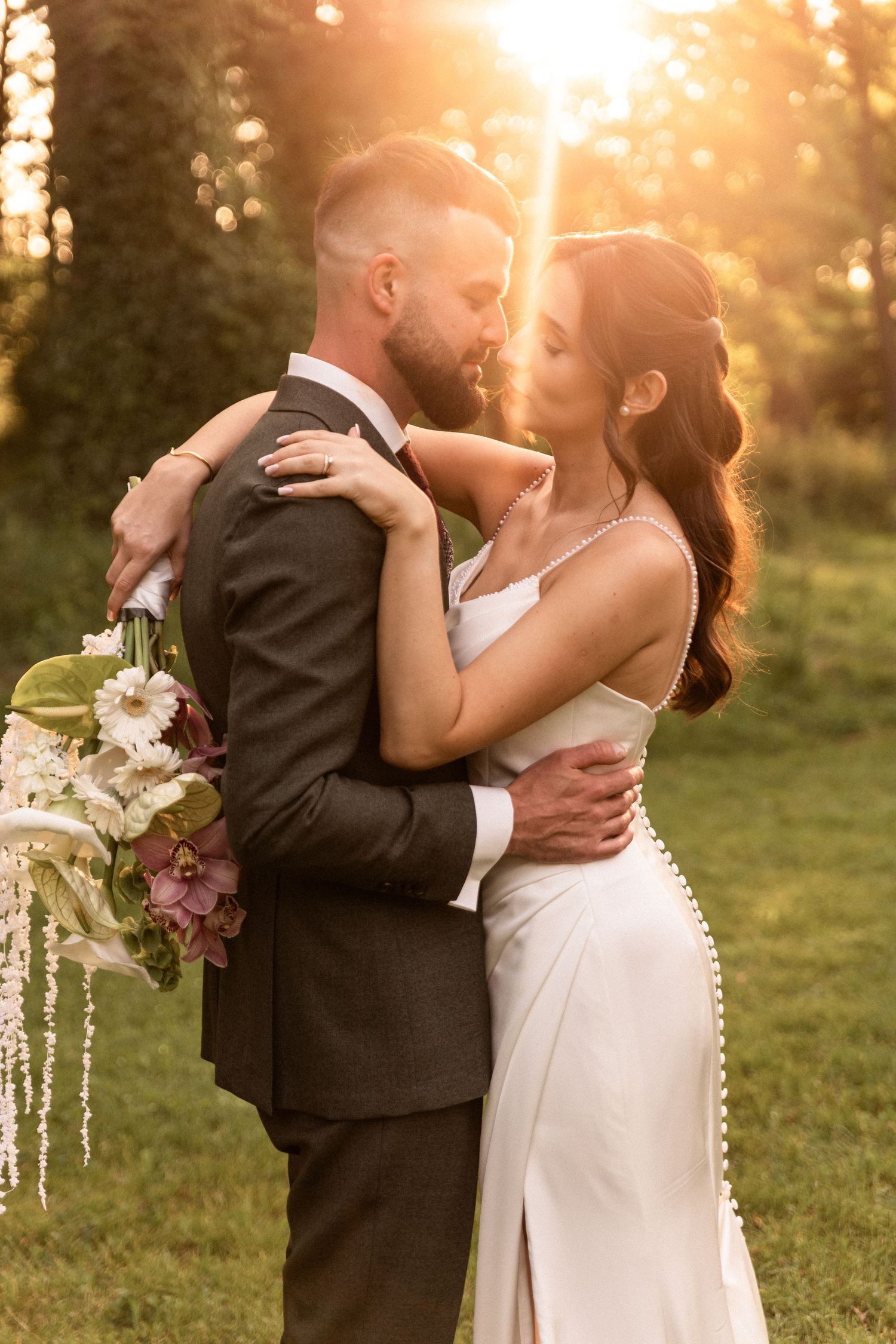 A romantic wedding couple embraces during golden hour sunset in a garden setting with a beautiful floral bouquet.
