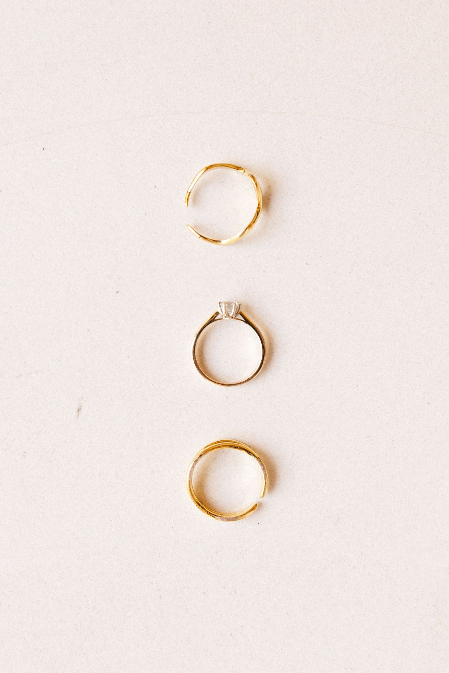 Three minimalist rings arranged vertically on white background, including one solitaire engagement ring and two gold bands.