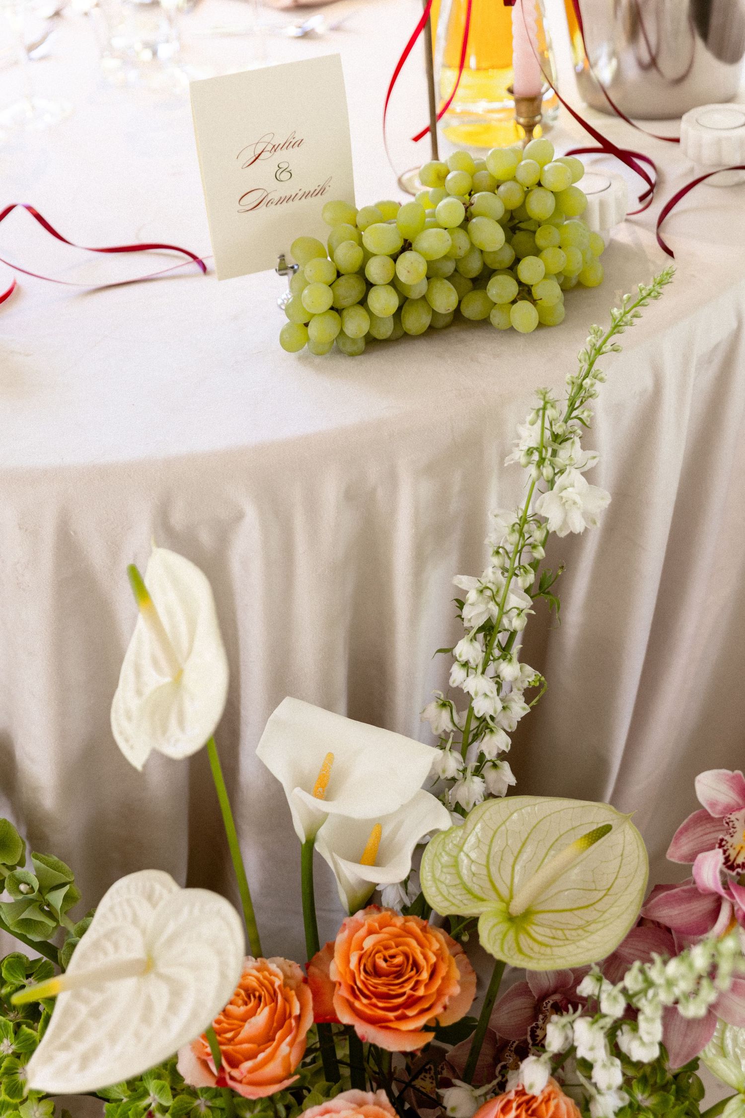 Elegant floral arrangement with white anthurium lilies, orange roses, grapes, and delicate snapdragon blooms on a tablecloth.