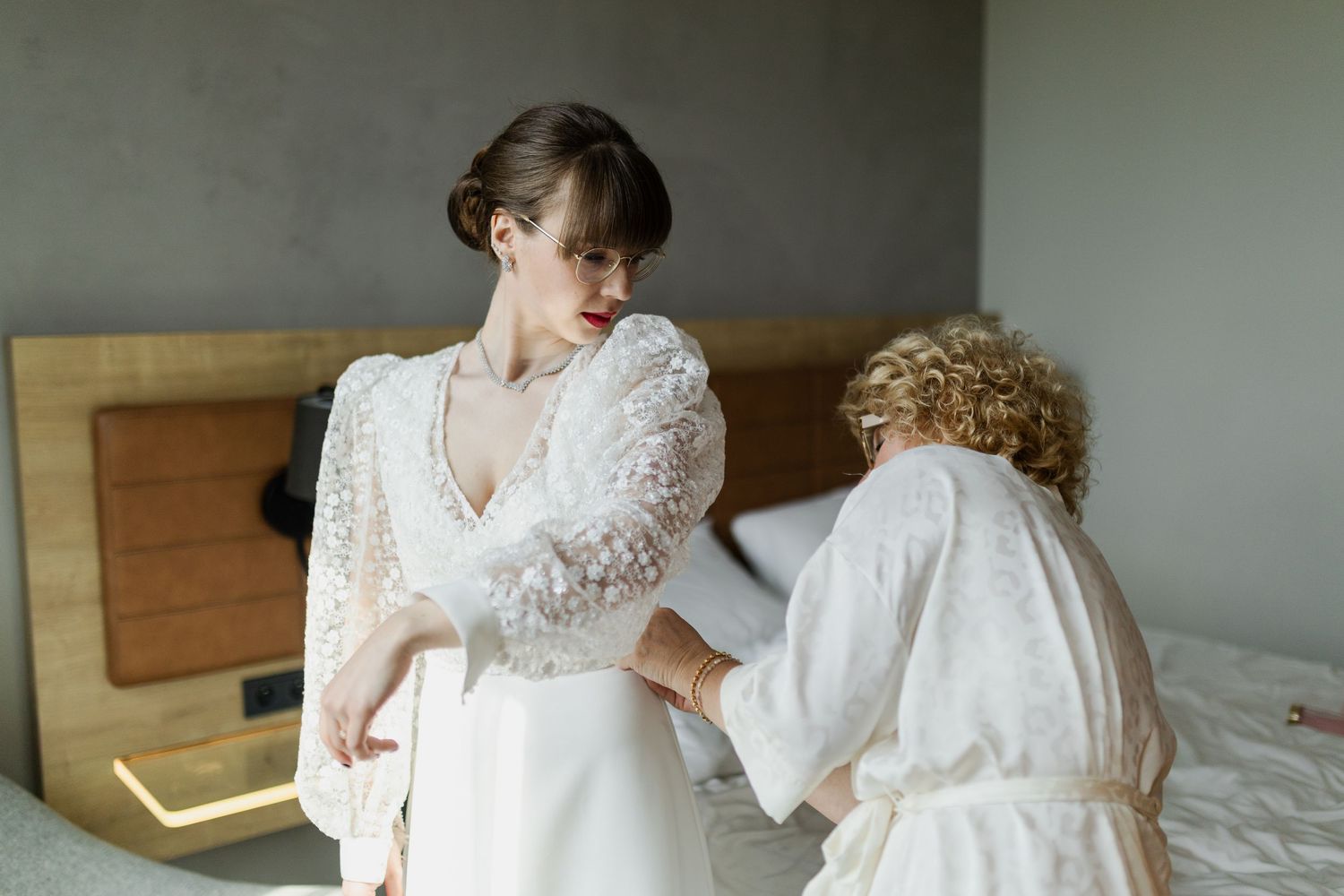 Two people in white lace dresses prepare for a special occasion in a bedroom setting.