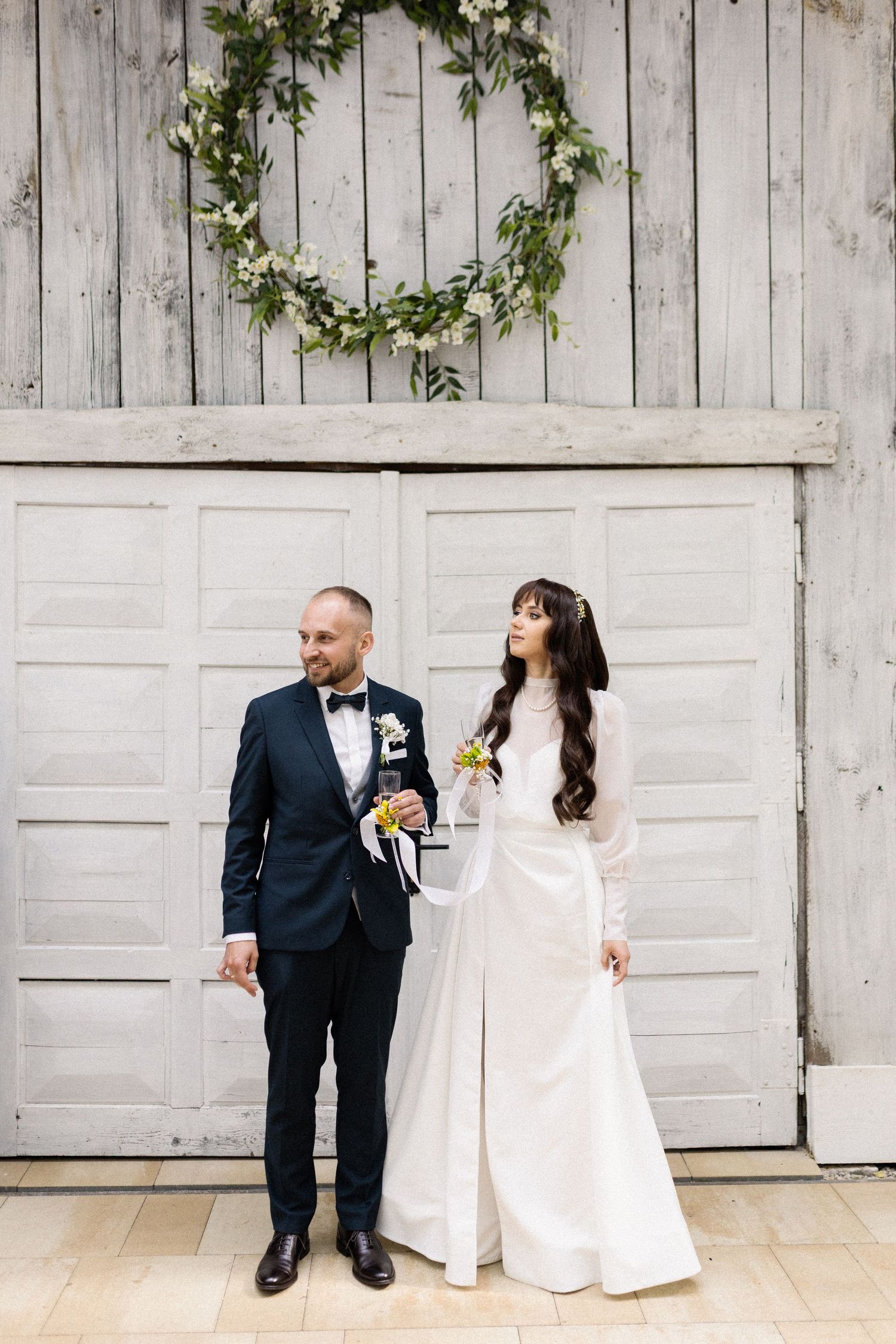 Wedding couple poses in front of rustic white barn doors decorated with a floral wreath.