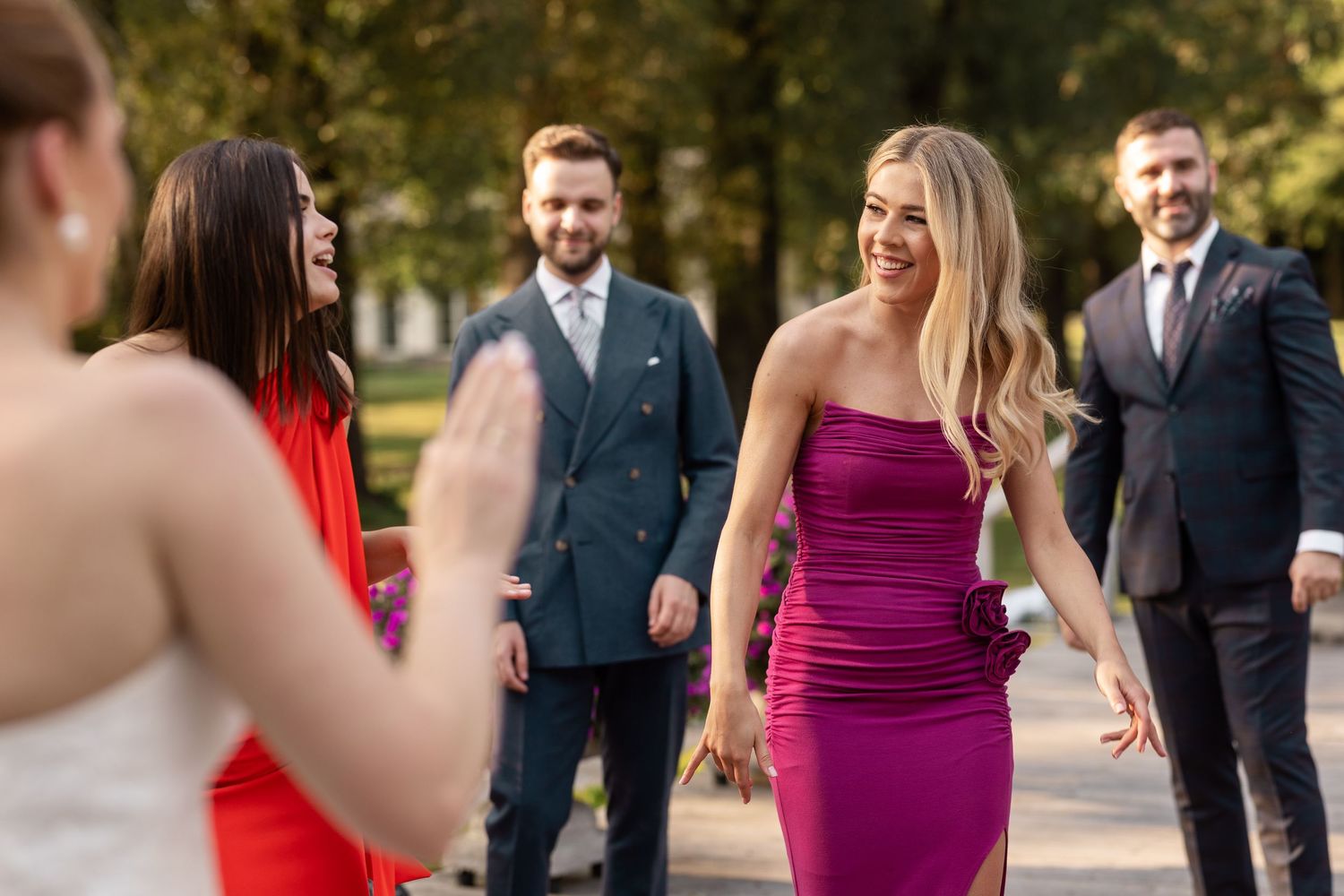 Wedding guests in formal attire laugh and socialize outdoors, with a woman in a vibrant magenta dress standing out.