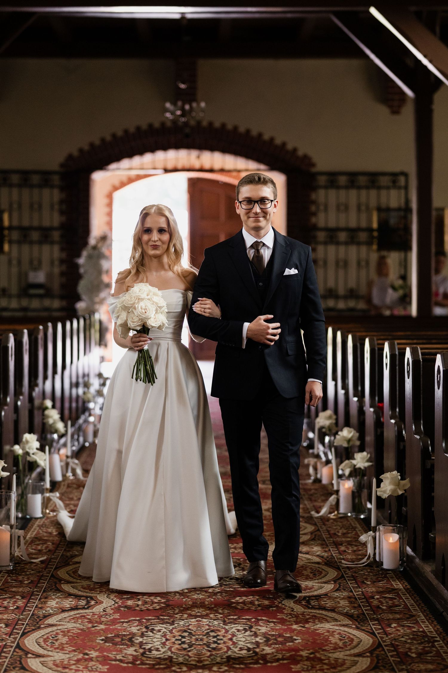 A couple walks down a church aisle decorated with candles and floral arrangements on a red patterned carpet.