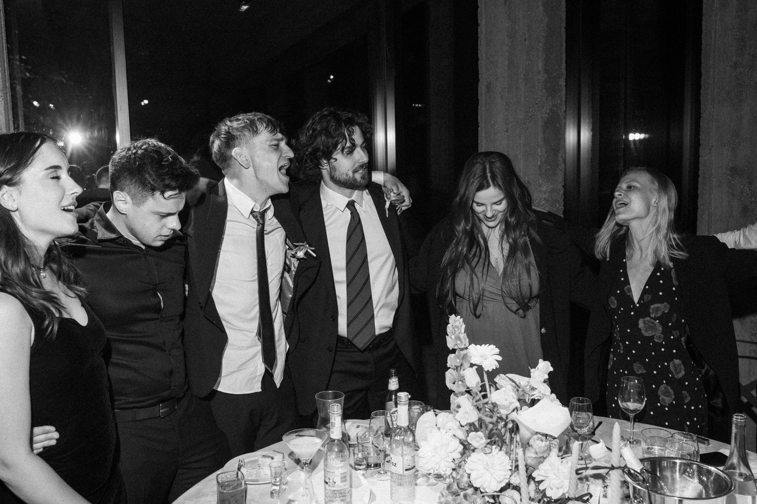 A candid black and white photo of a group laughing together at an elegant dinner party with table settings and flowers.