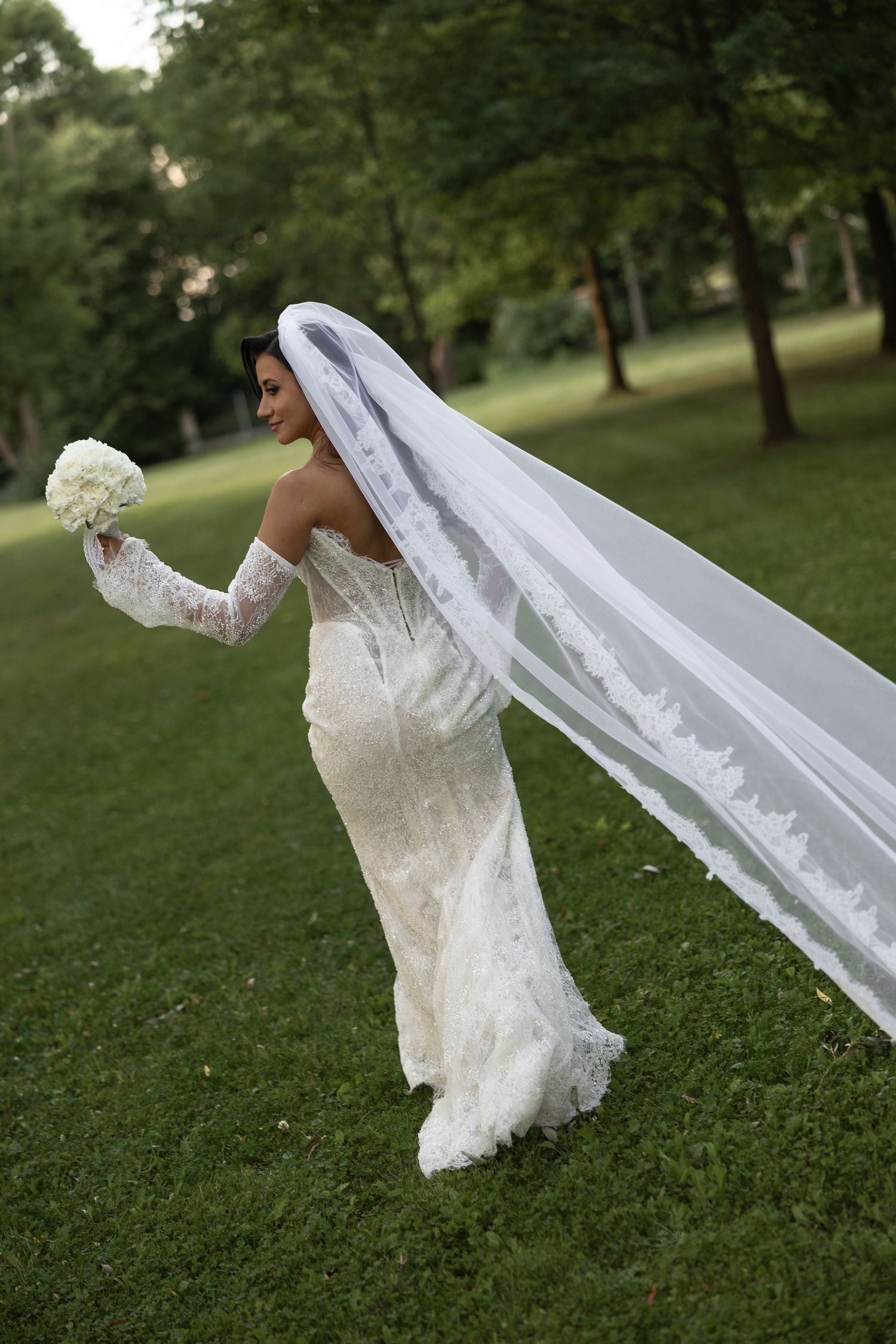 Bride in lace wedding gown with long flowing veil holds white bouquet while posing in lush green park.